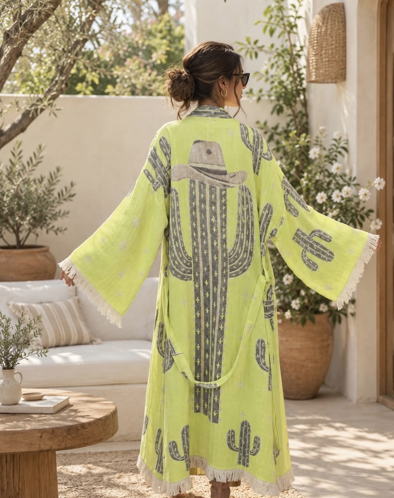 Person wearing a bright yellow dress with cactus and hat patterns, standing outdoors with plants and a white wall behind.