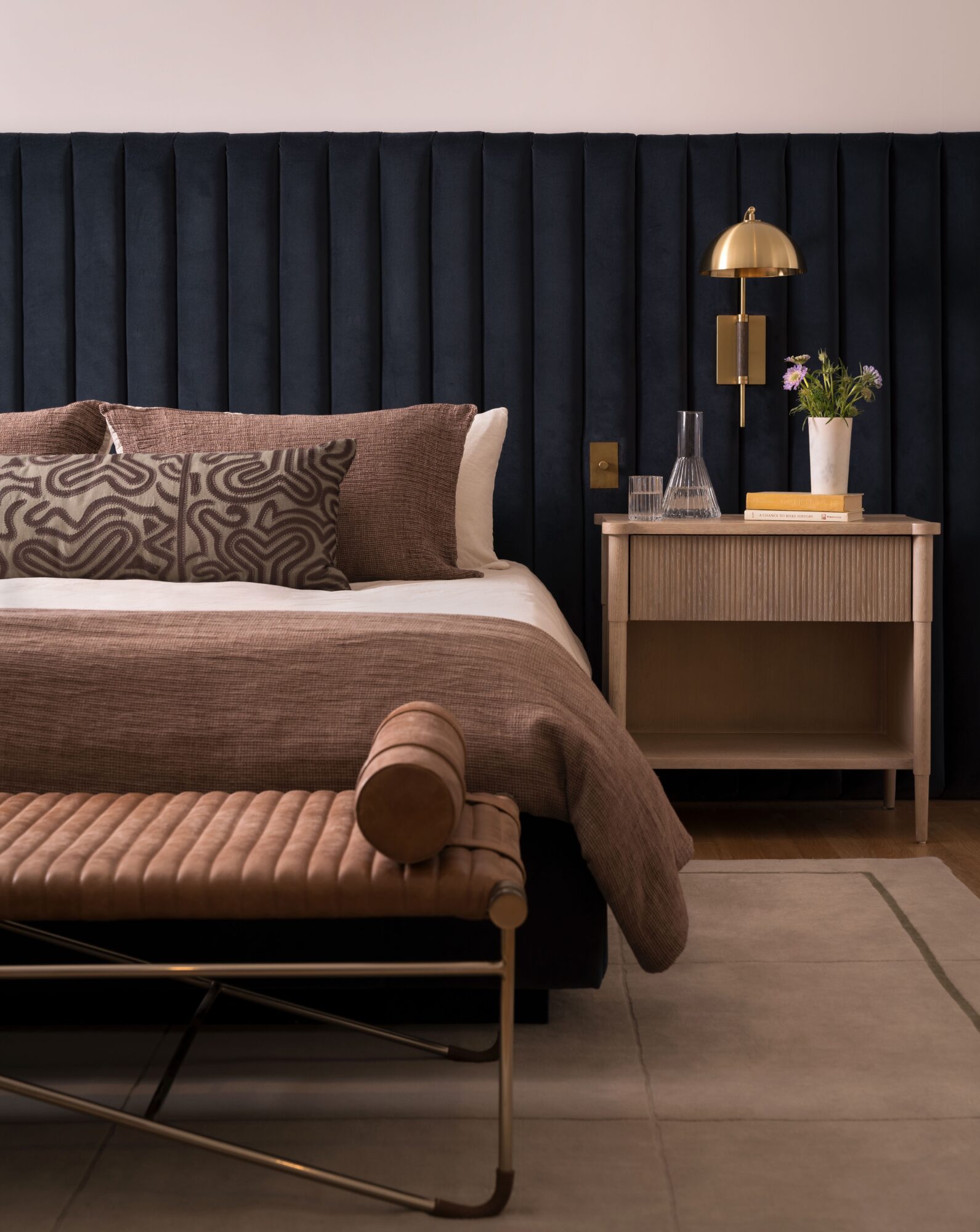 Bedroom with a bed, patterned pillows, a cushioned bench, and a bedside table with a lamp and plant.