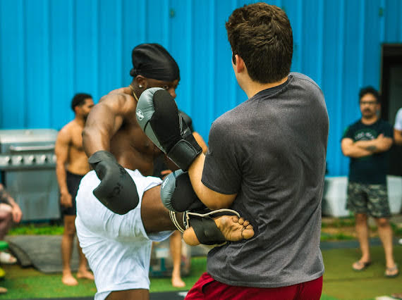 Two people practice a martial arts hold on grass, with others standing nearby.