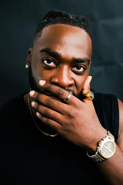 Portrait of a person wearing jewelry, with one hand covering their mouth against a dark background.