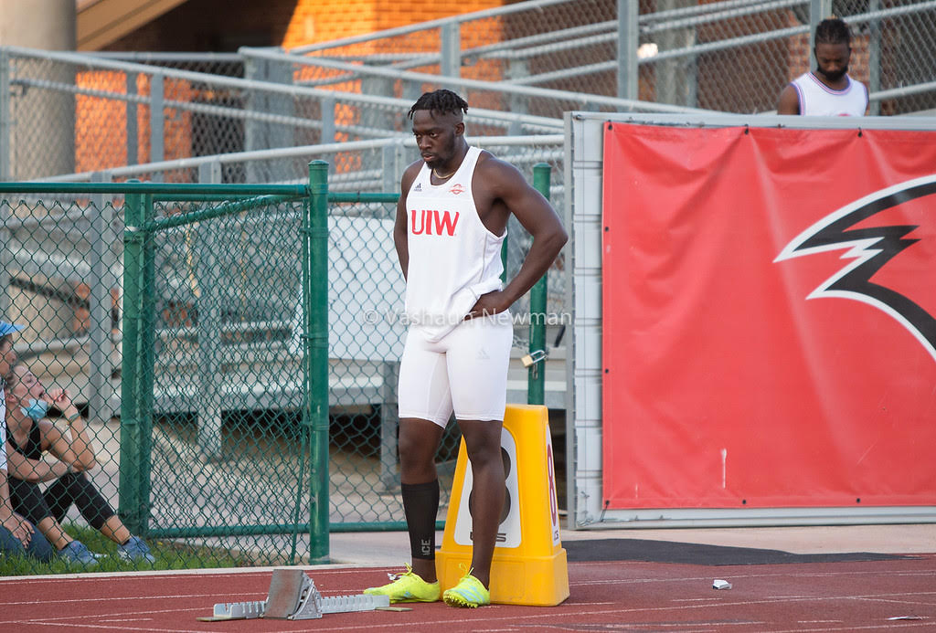 Athlete in a white uniform stands on a track beside a fence and red team banner.