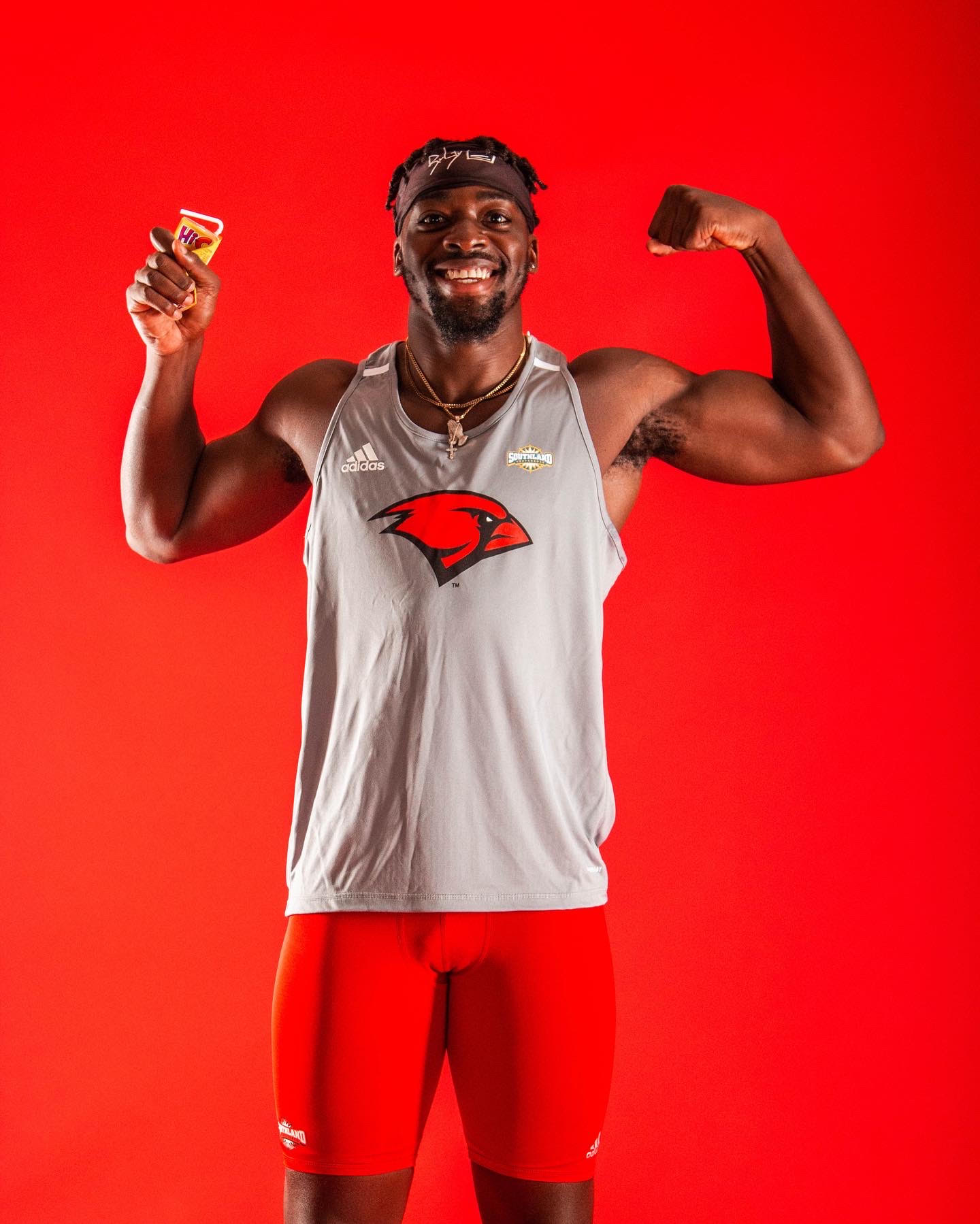 Person in a gray basketball jersey flexes one arm and holds a small item against a red background.