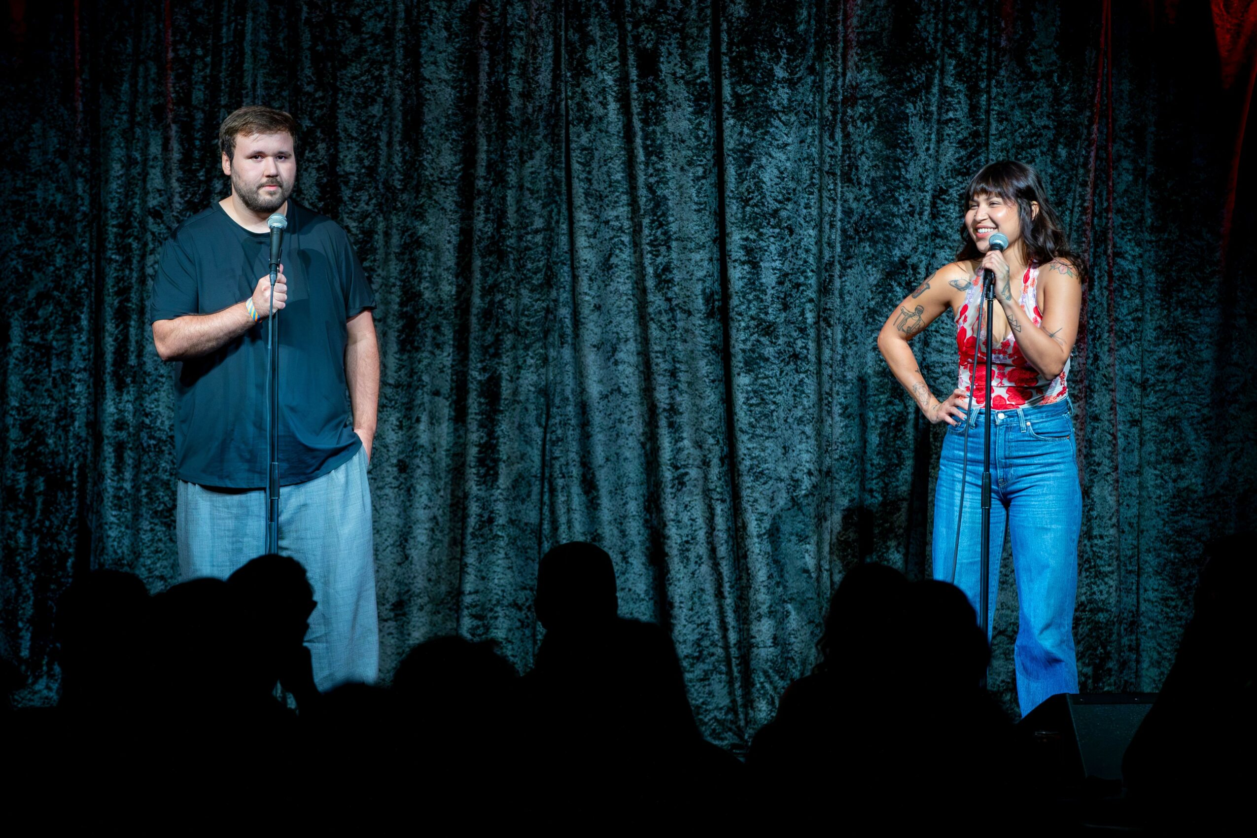 Two people stand on stage with a dark curtain background, holding microphones, engaging with an audience.