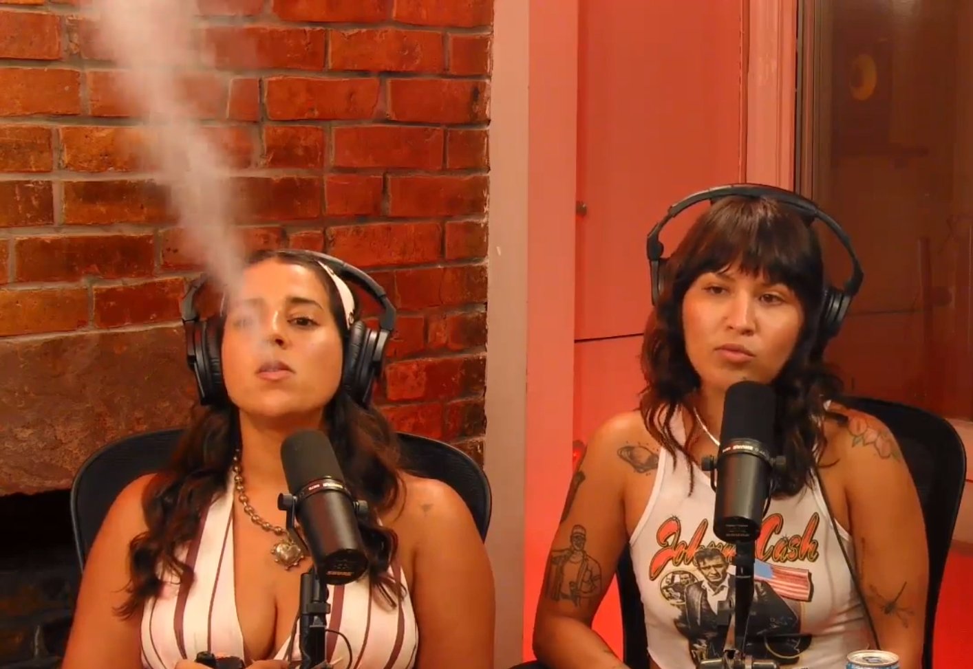Two women with headphones sit in front of microphones, one is exhaling smoke, in a studio with brick wall background.