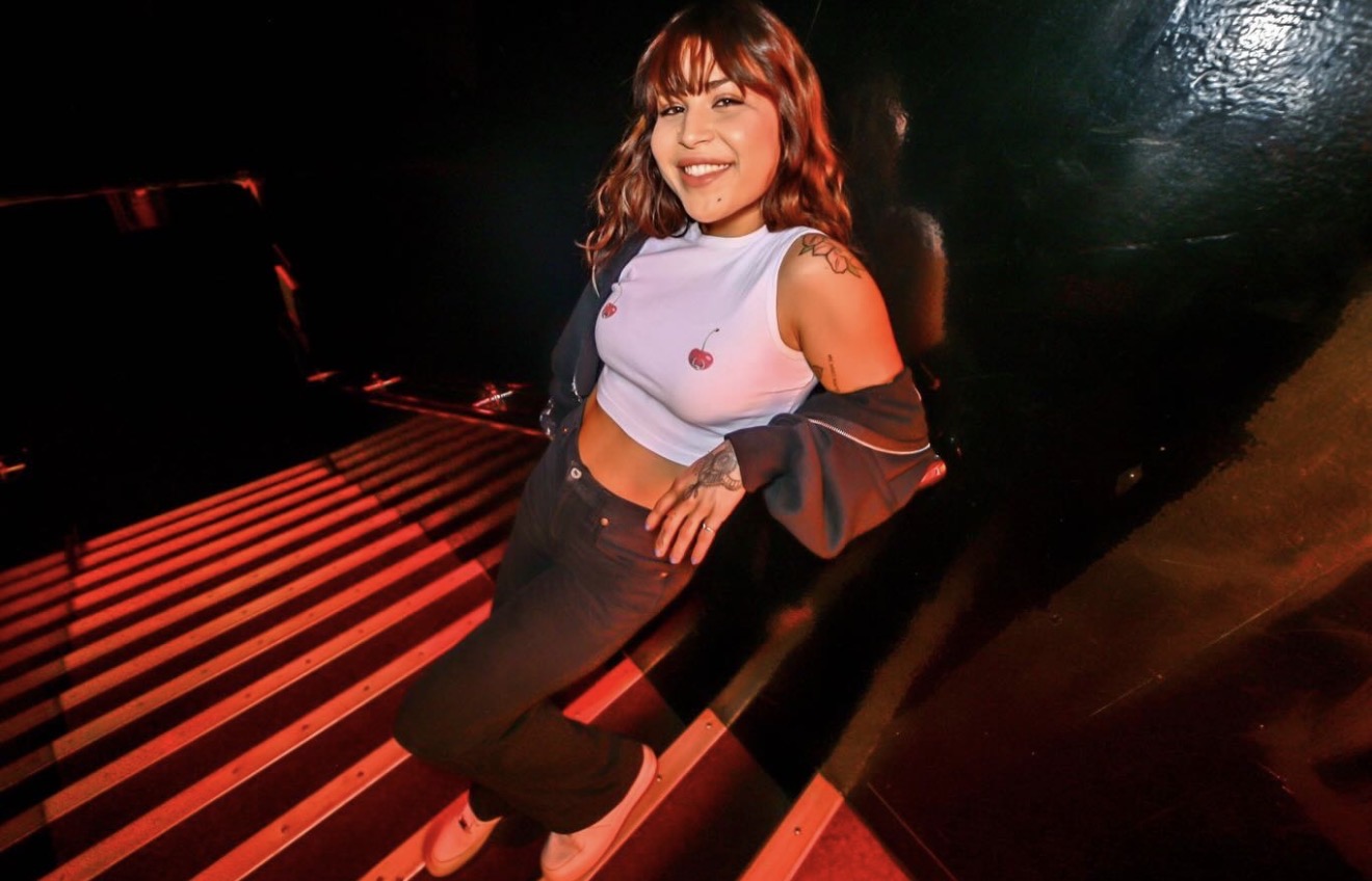 Person with shoulder-length hair smiling, sitting on a striped surface, wearing a white top with cut-out shoulders and dark pants.