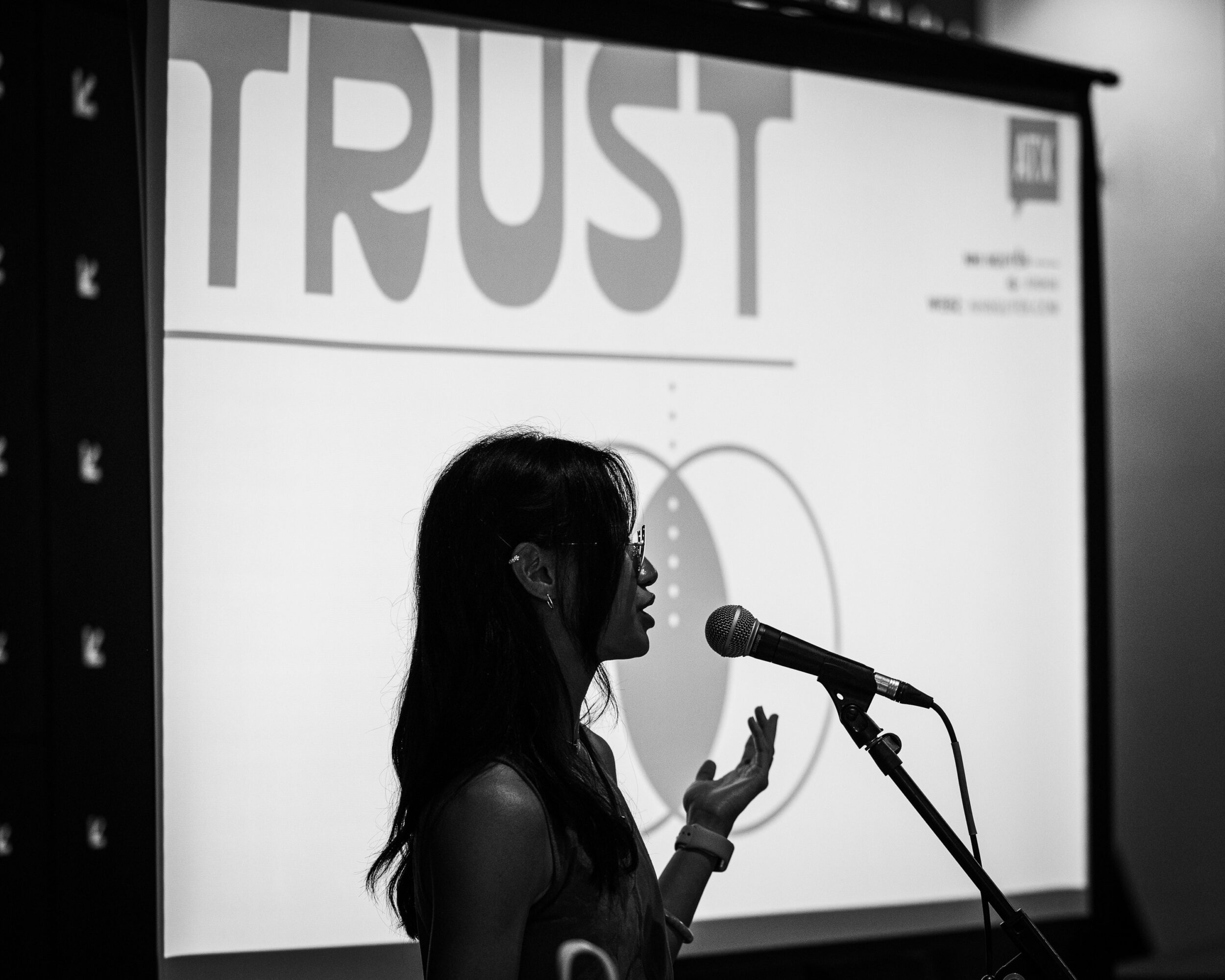A person speaks into a microphone in front of a projected slide with the word “TRUST.”