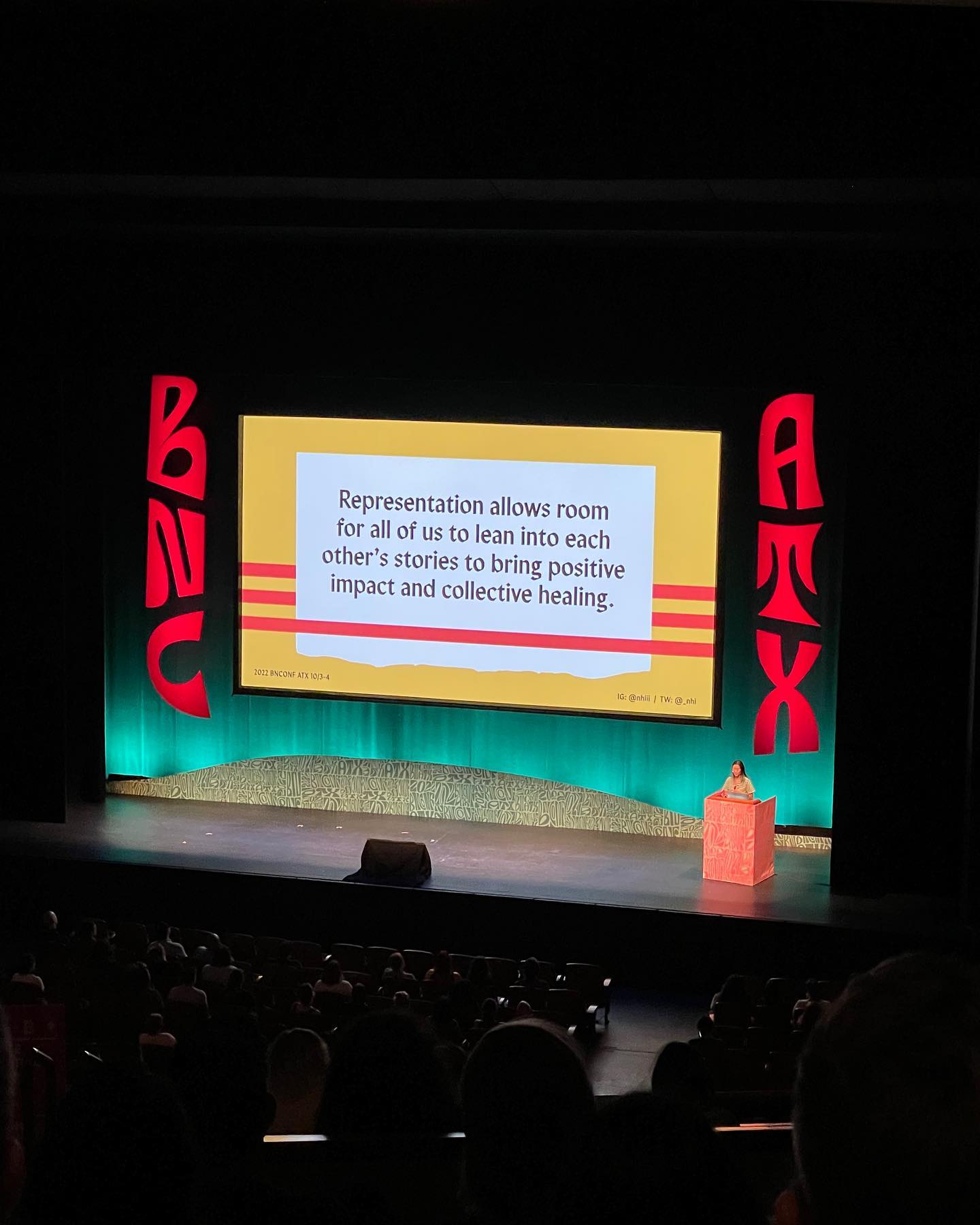 Stage screen displays a message about representation, with red lettering, colored lights, and audience seating in front.