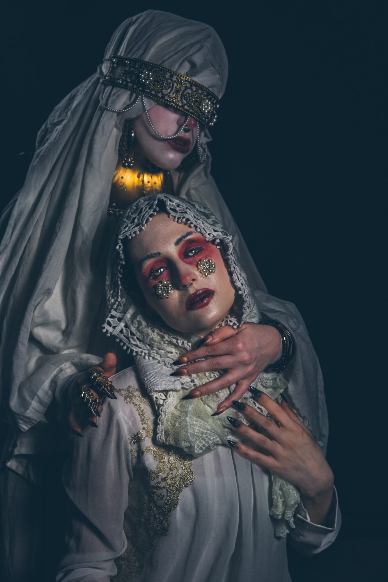 A veiled person stands behind another person wearing jeweled face decorations and holding a small object.
