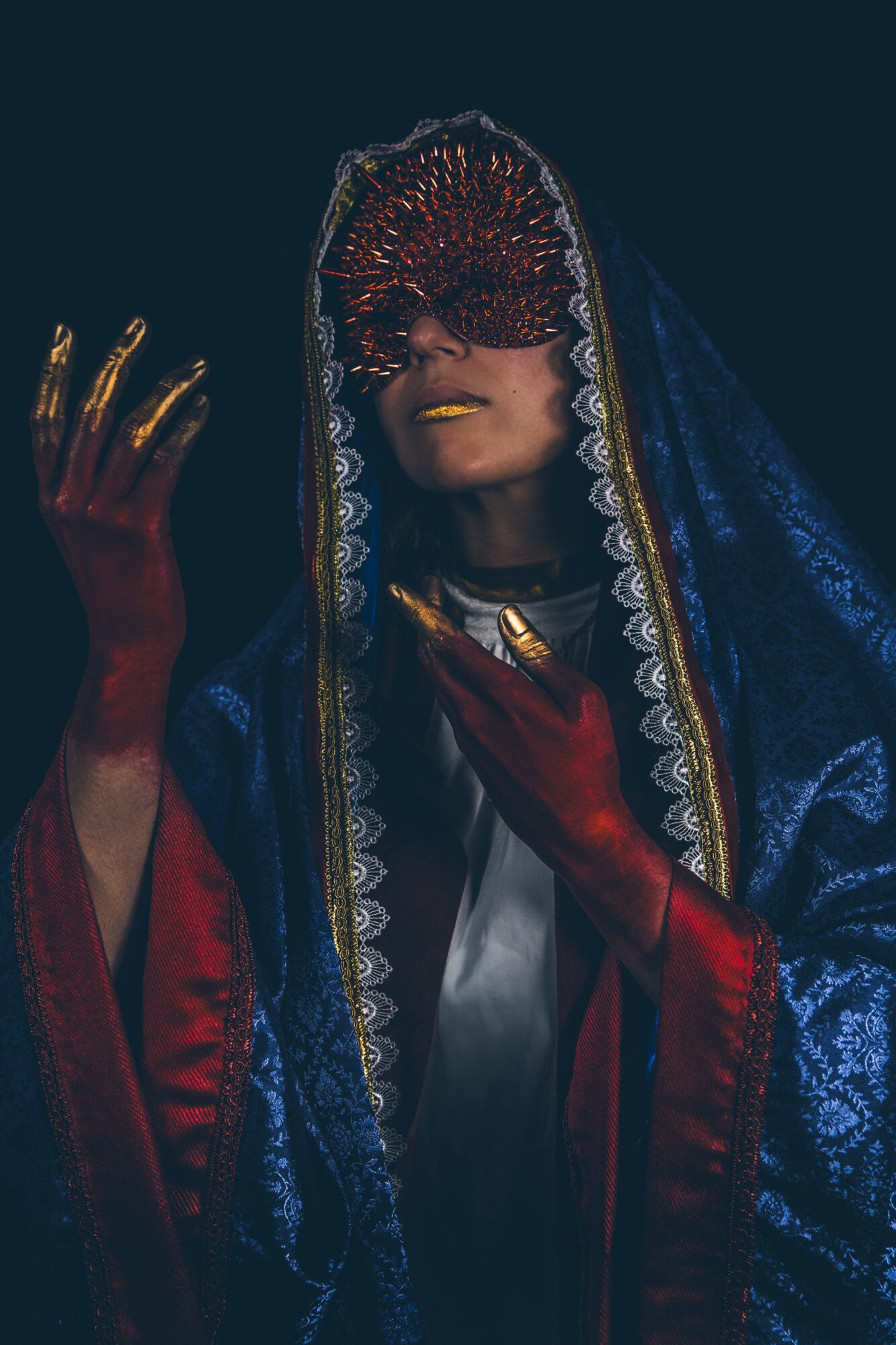 Person wearing an ornate dark blue veil and red garments, raising one hand against a dark background.