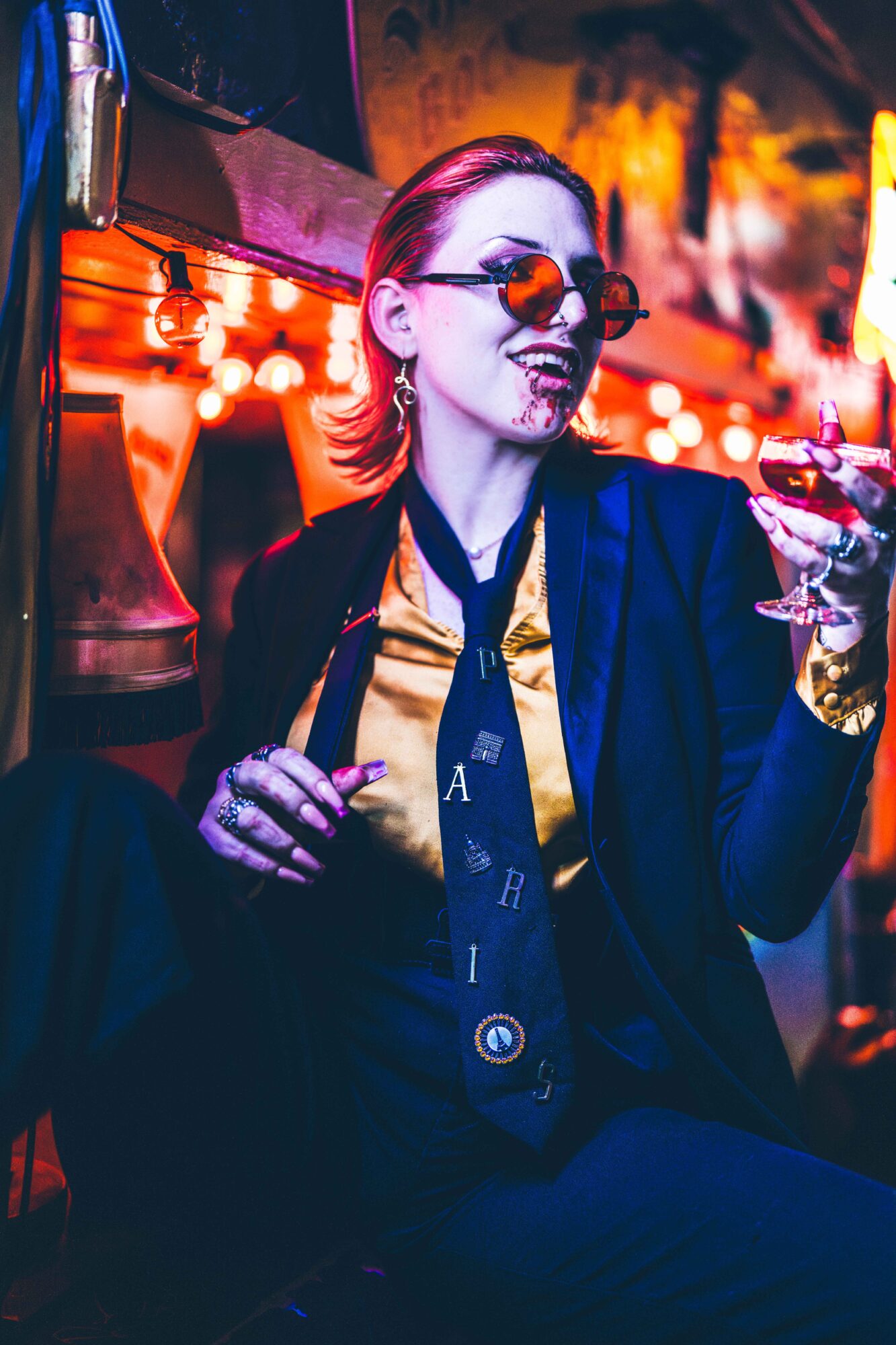 Person wearing sunglasses and a dark suit holds a cigarette and a drink in a warmly lit bar.