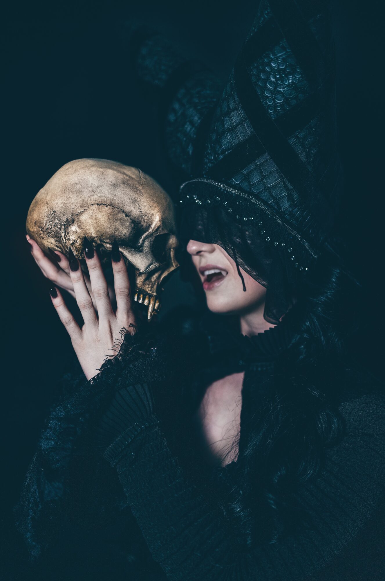 A person in a dark hood holds a human skull near their face.