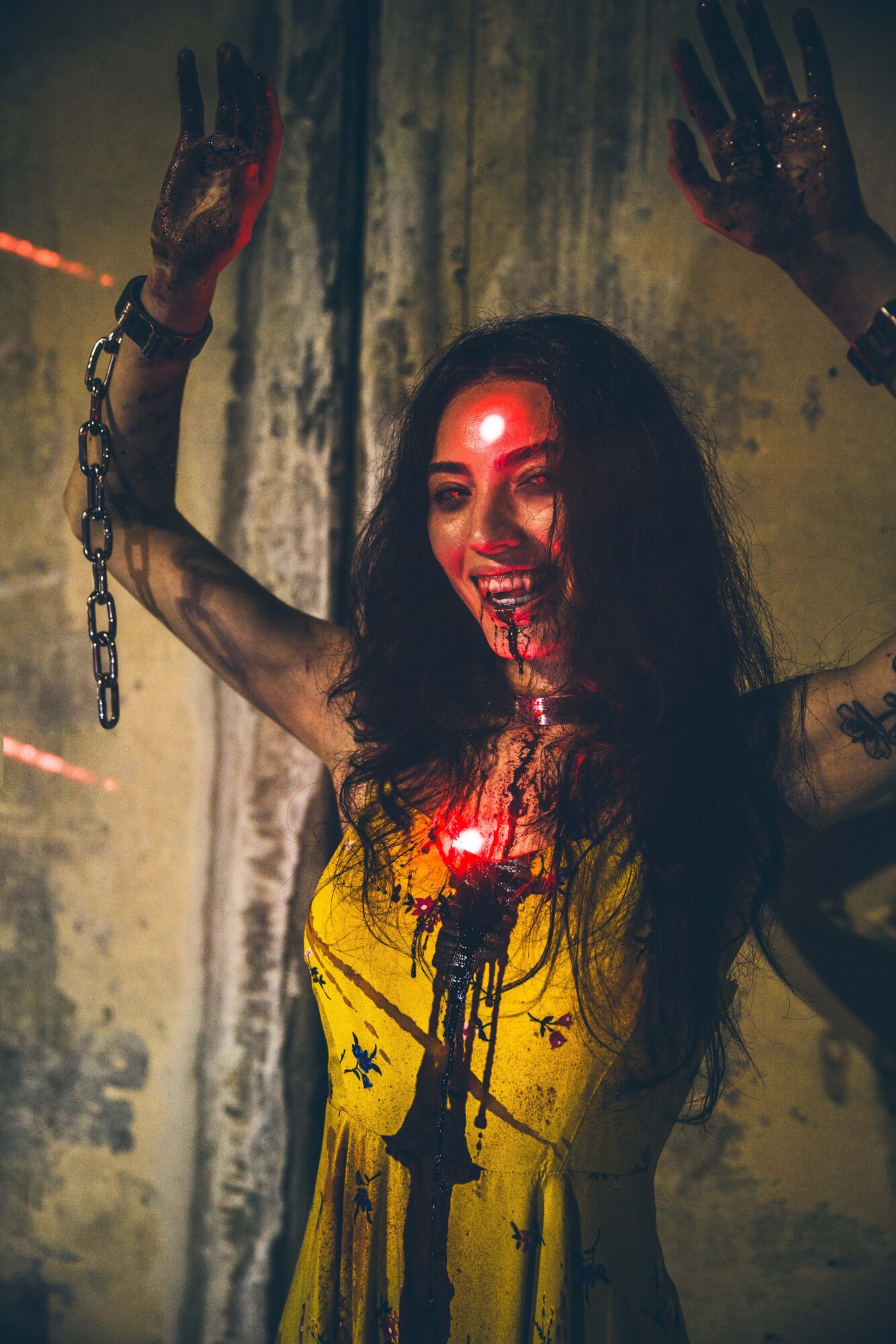 Person with long dark hair in a yellow dress raises both hands against a stained wall; chains and red lights are visible.