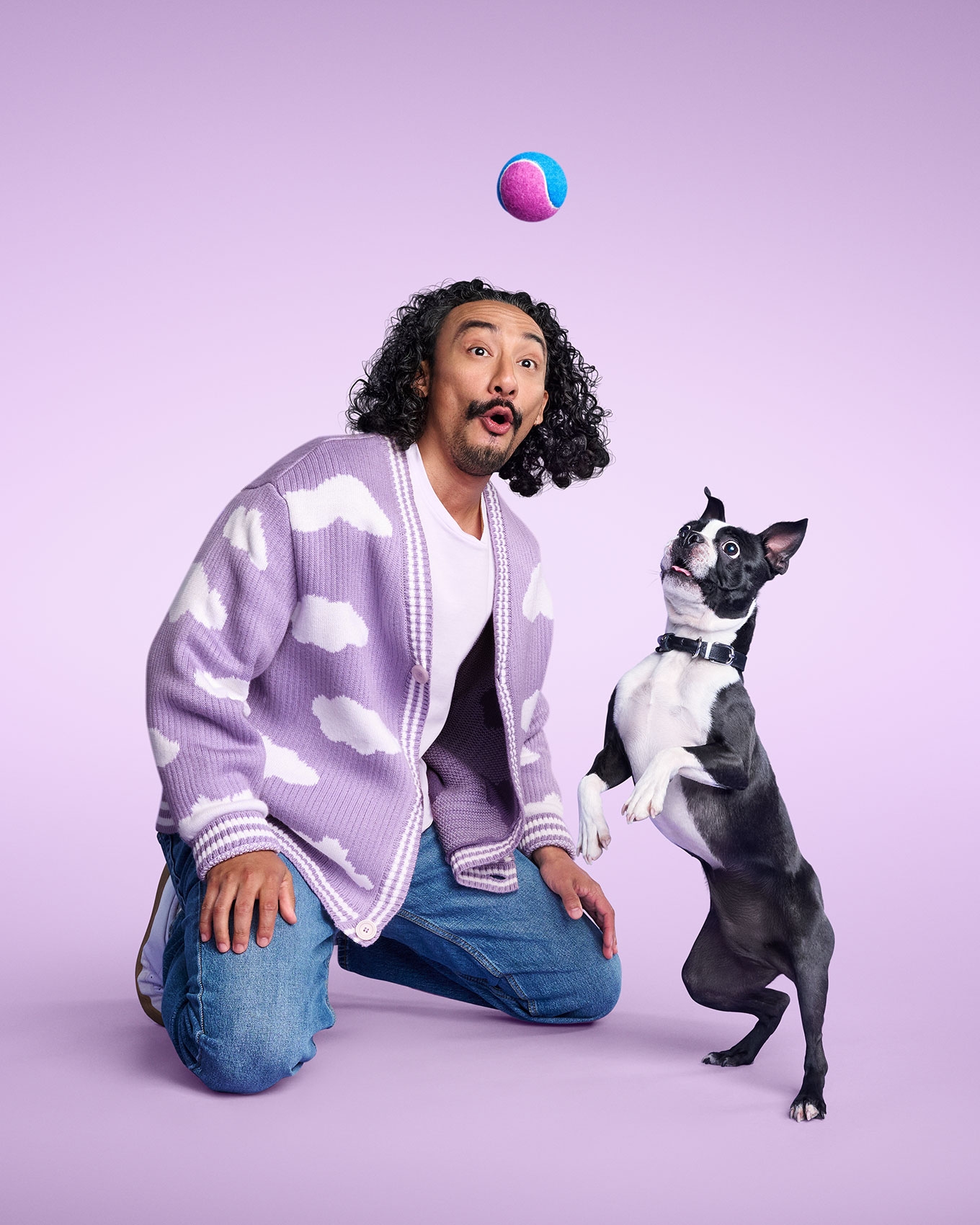 A person kneels beside a dog looking up at a small ball against a lavender background.