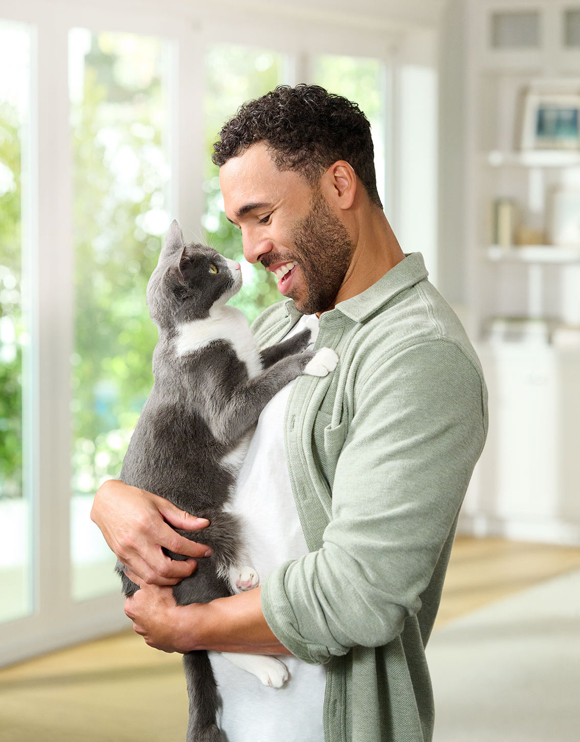 A man holds a gray-and-white cat near a bright window.