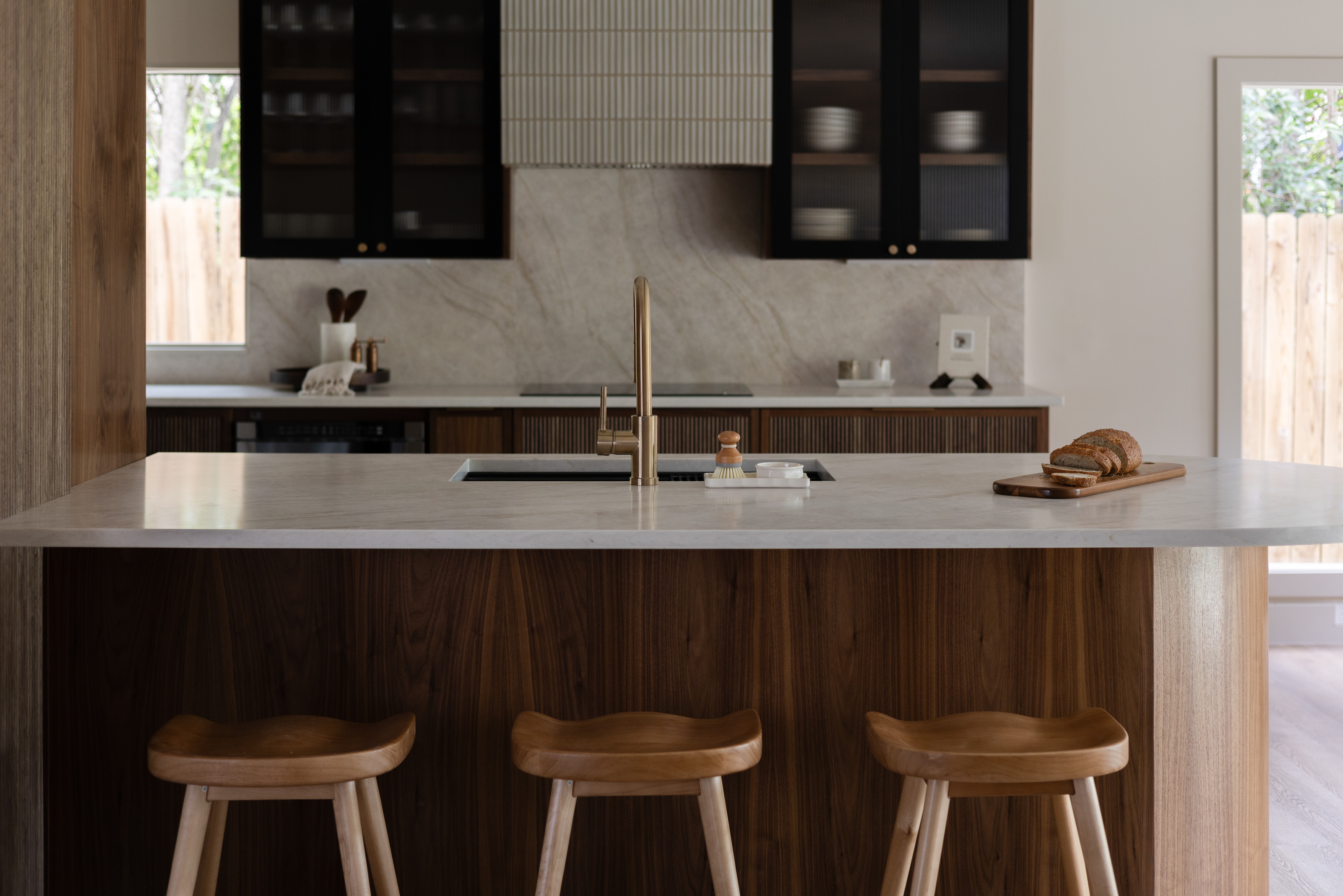 Modern kitchen with a central island, three wooden stools, dark cabinets, and windows on both sides.