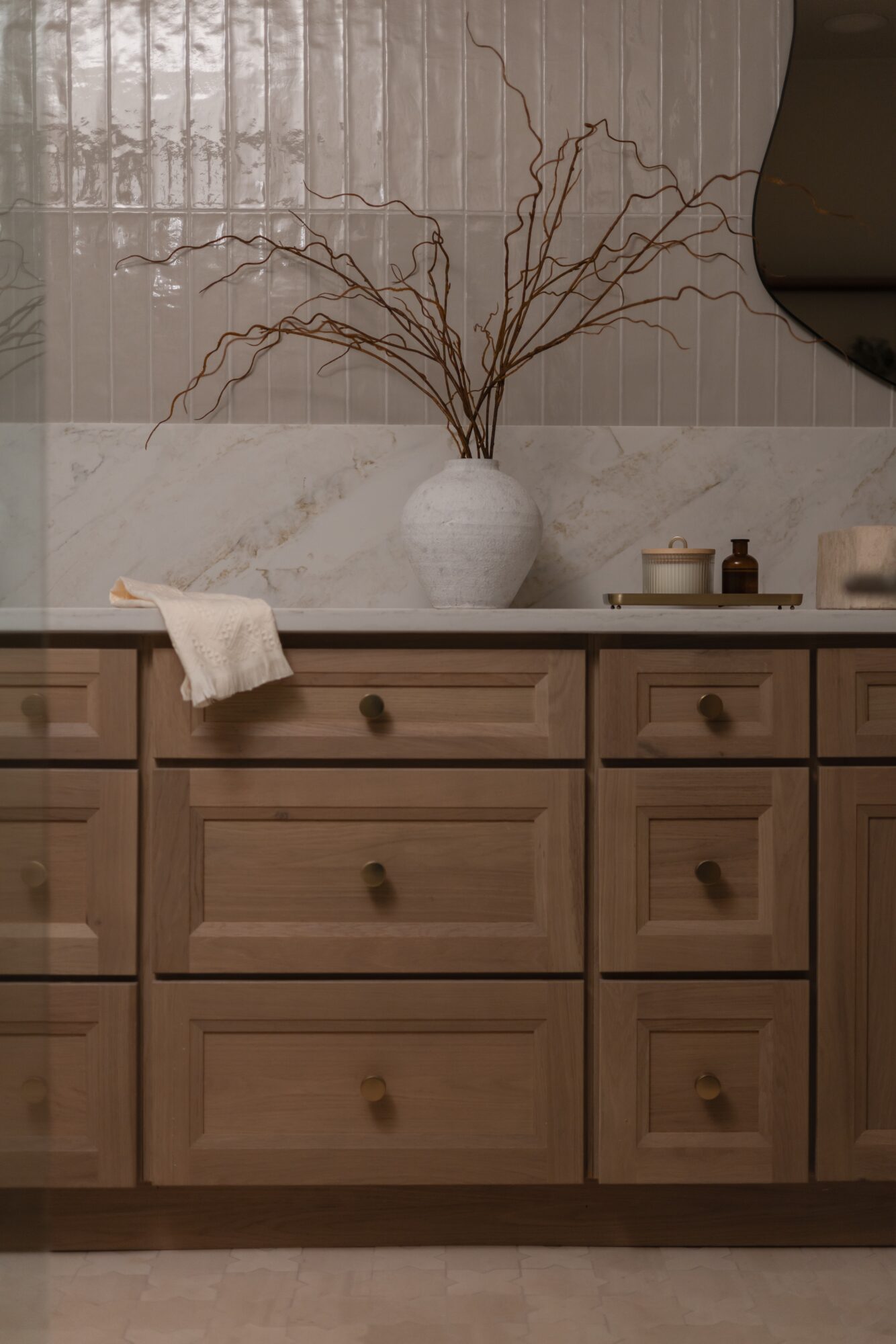 A white vase with bare branches sits on a bathroom vanity beneath a window, beside small containers and a sink.