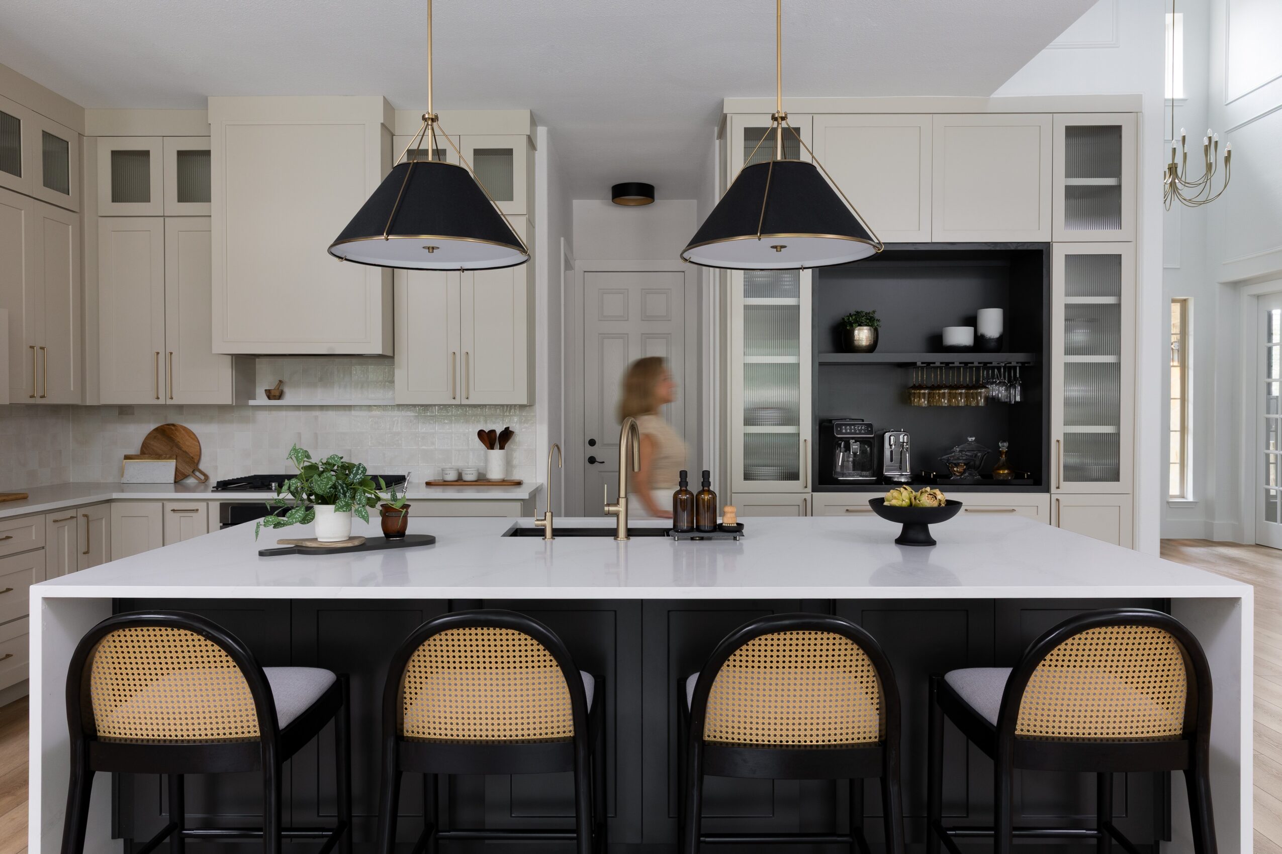 Bright kitchen with a large island, four stools, hanging lights, and a person standing by the counter.
