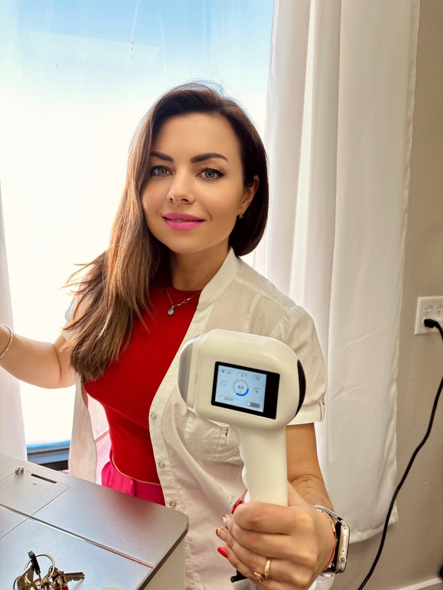 A person in a white coat holds a handheld device beside a table and bright window.