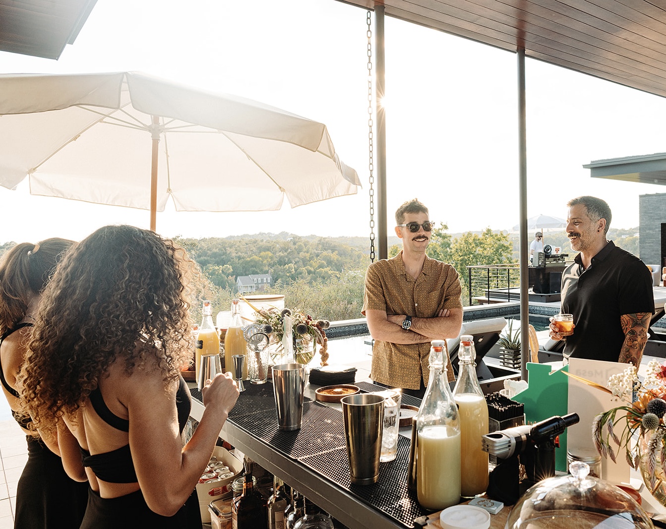 People gather around an outdoor bar beneath umbrellas, with drinks and bottles on the counter.