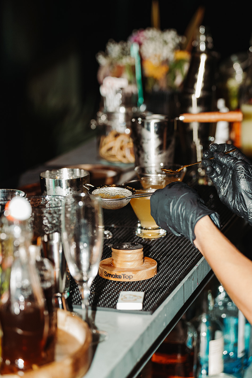 Cocktail bar counter with glassware, bottles, a drink, and gloved hands preparing a beverage.