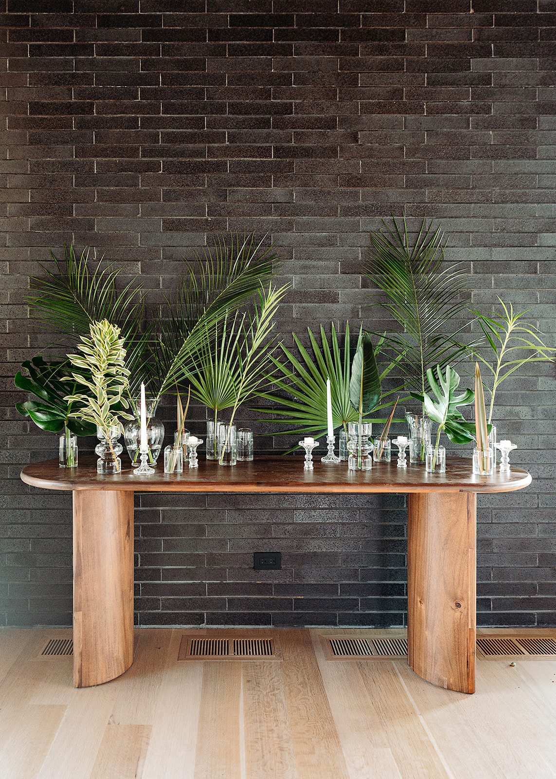 Wooden console table with glass vases and tall green plants against a dark brick wall.