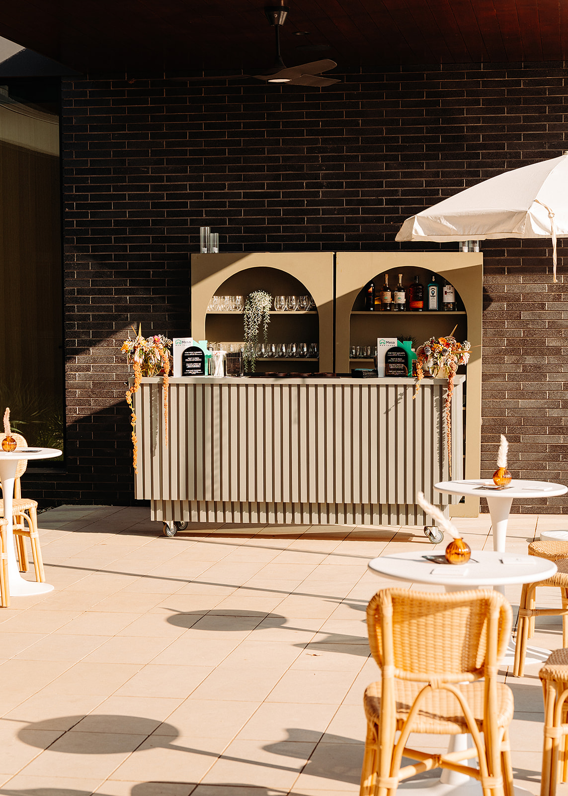 Outdoor bar with a striped counter, stools, round tables, and a large umbrella against a brick wall.