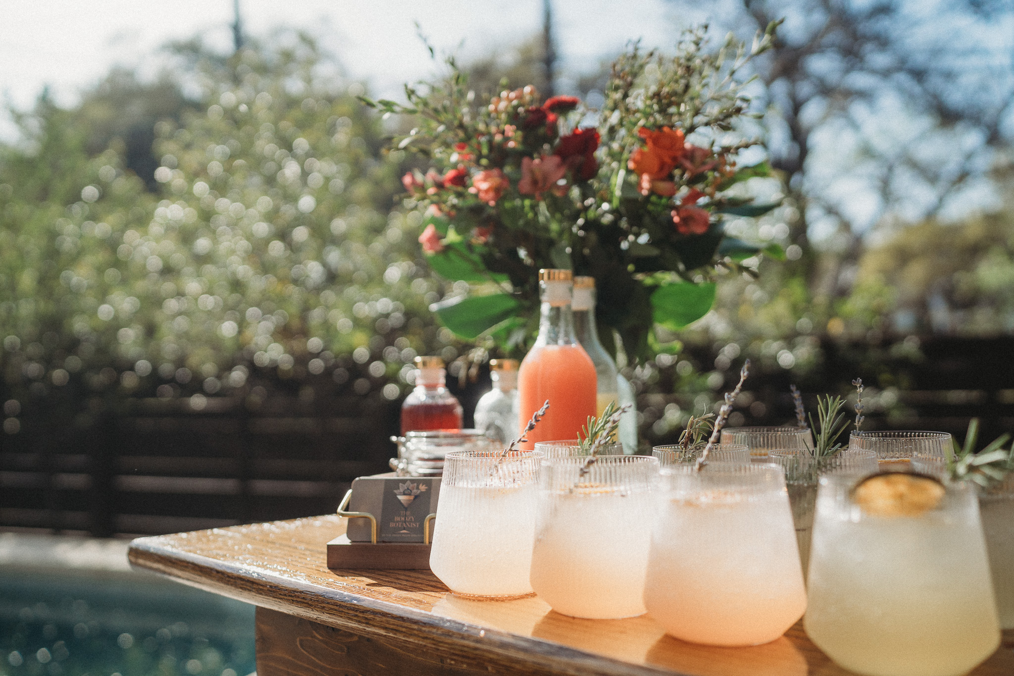 A table with colorful drinks and a flower arrangement outdoors, with a pool and trees in the background.