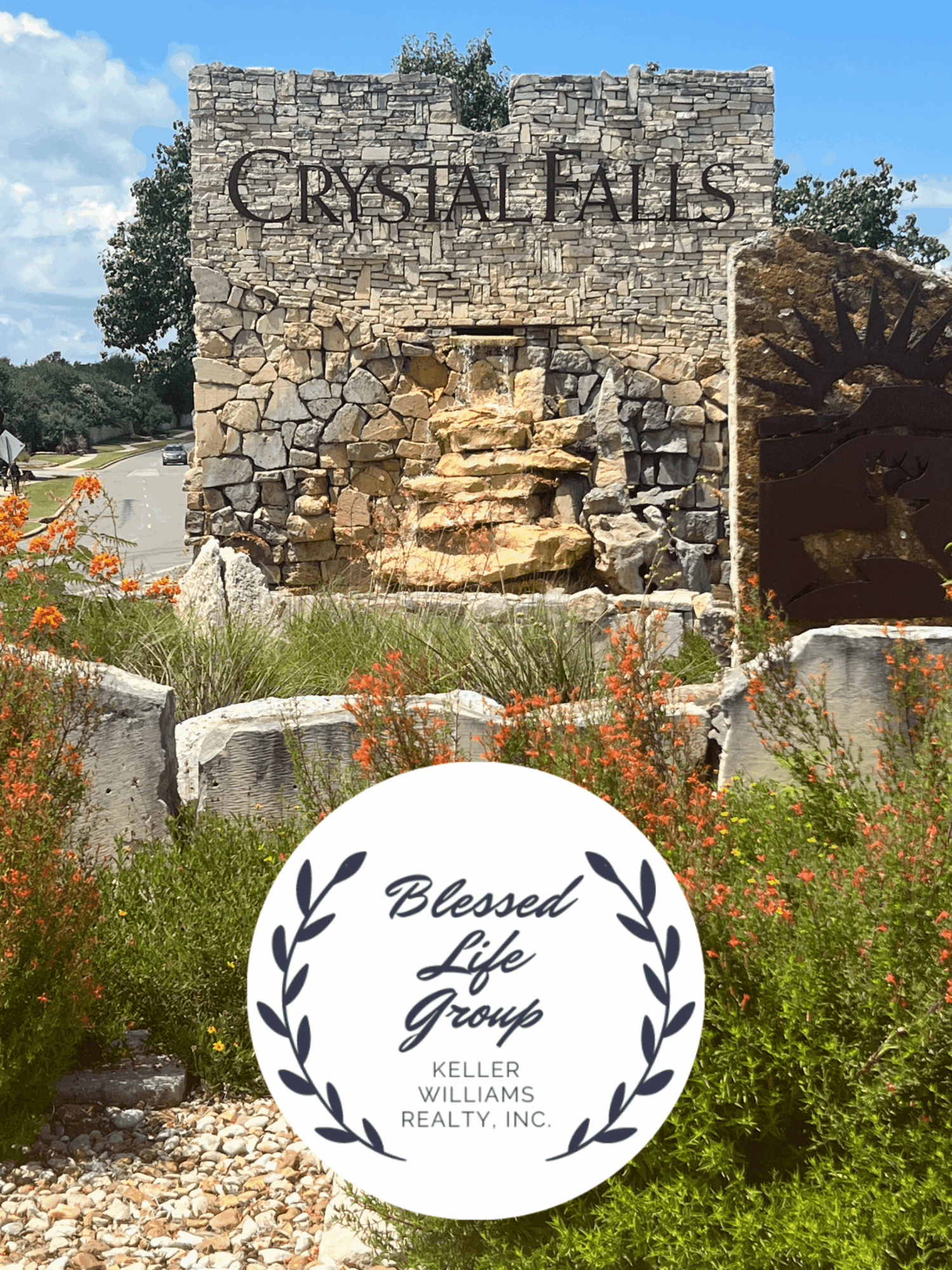 Stone entrance sign reading “Crystal Falls,” with a small waterfall, flowers, and a circular Blessed Life Group logo.