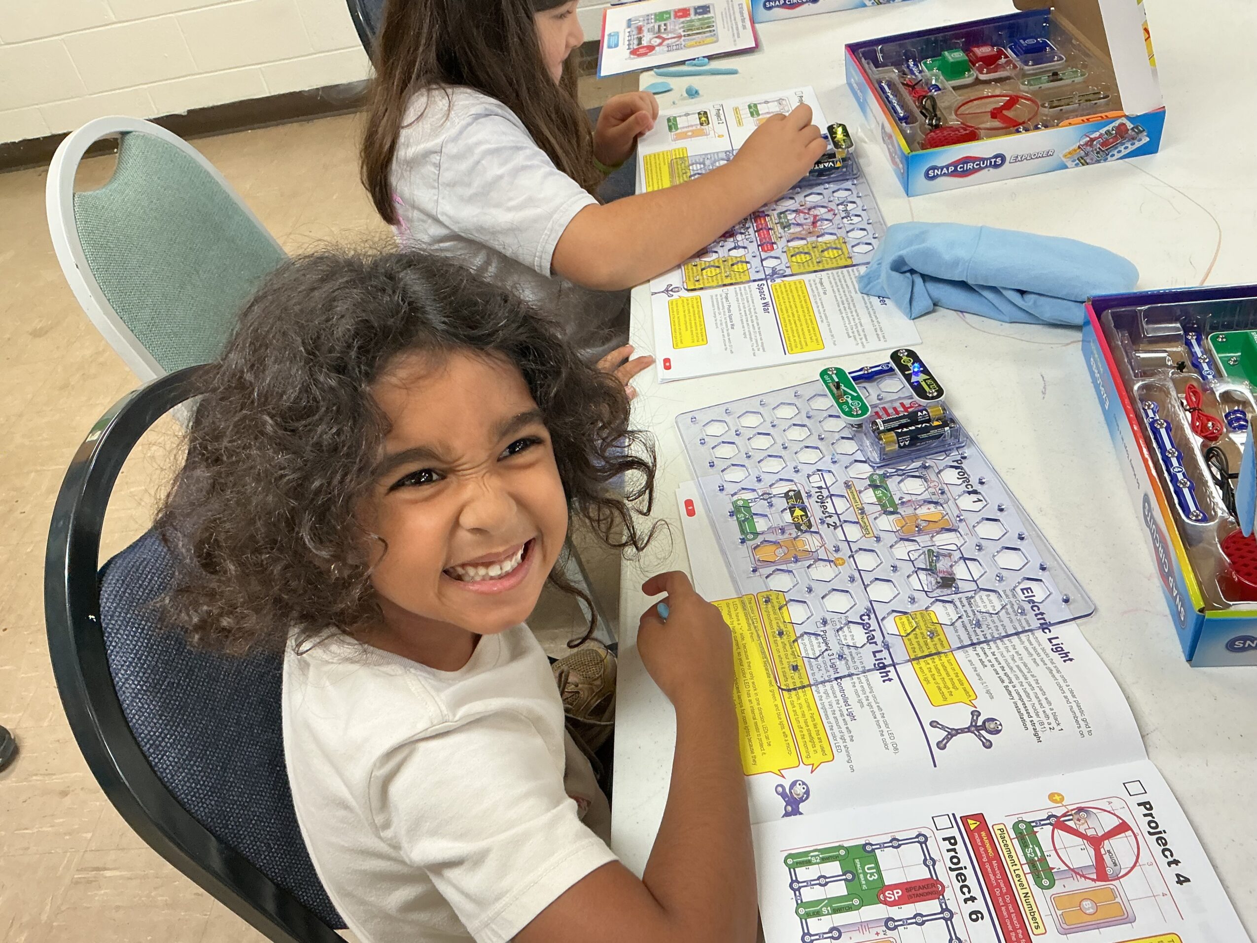 Two children sit at a table with colorful activity sheets and craft supplies; one smiles toward the camera.