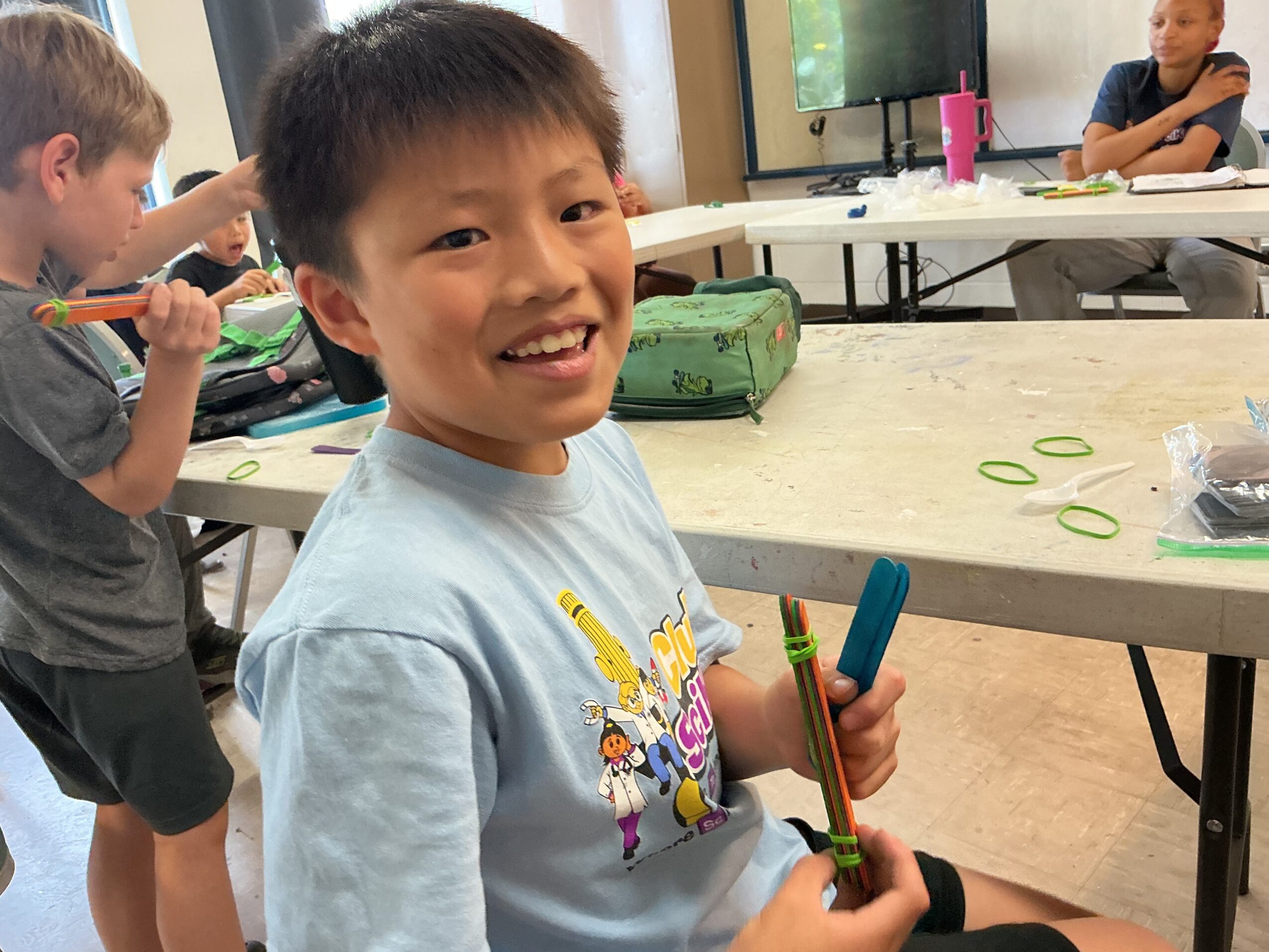 A child holding colorful craft sticks at a table with green craft shapes; other people sit nearby.