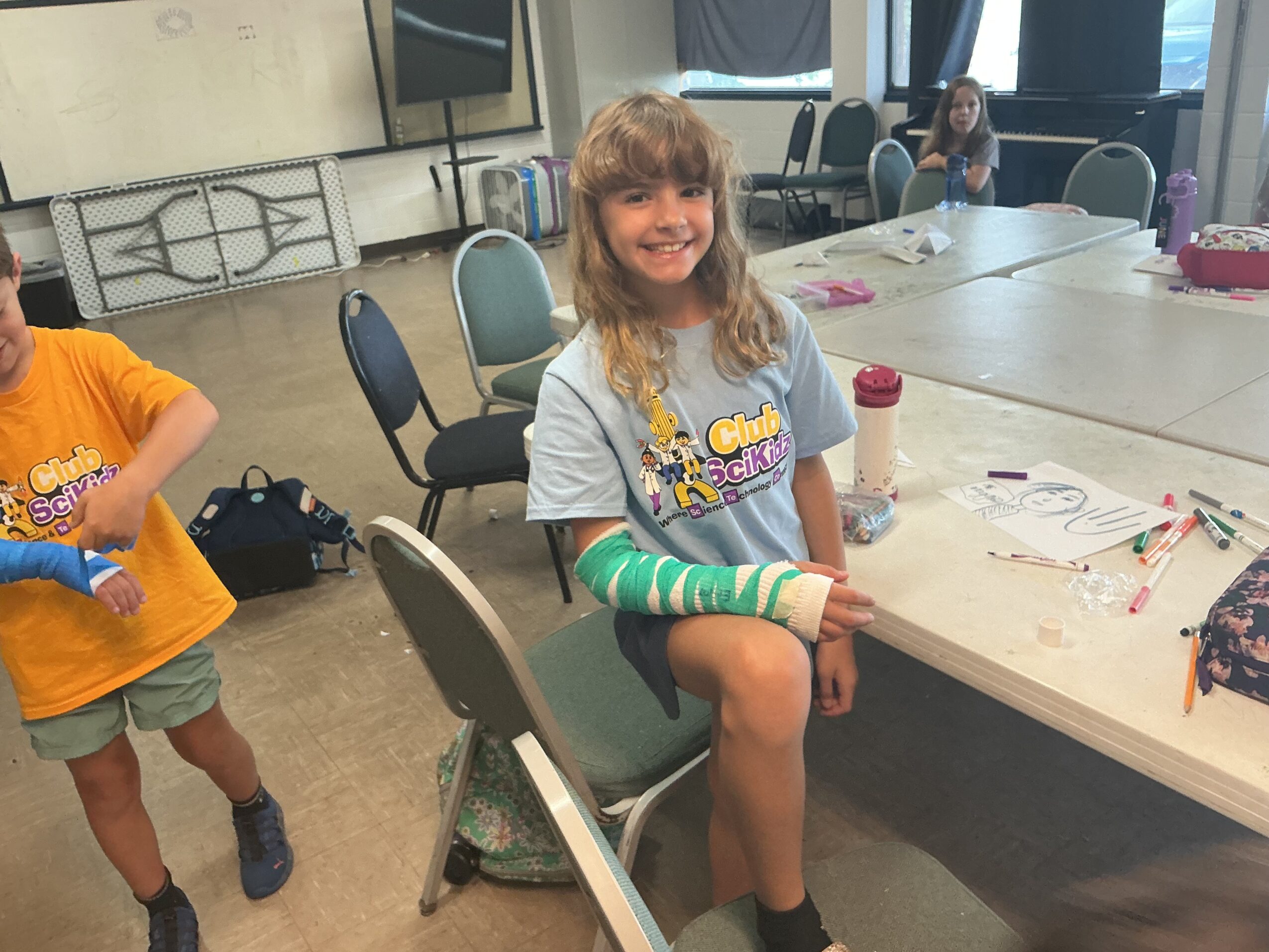 A seated child with a green arm cast smiles at a craft table, while other people work nearby.