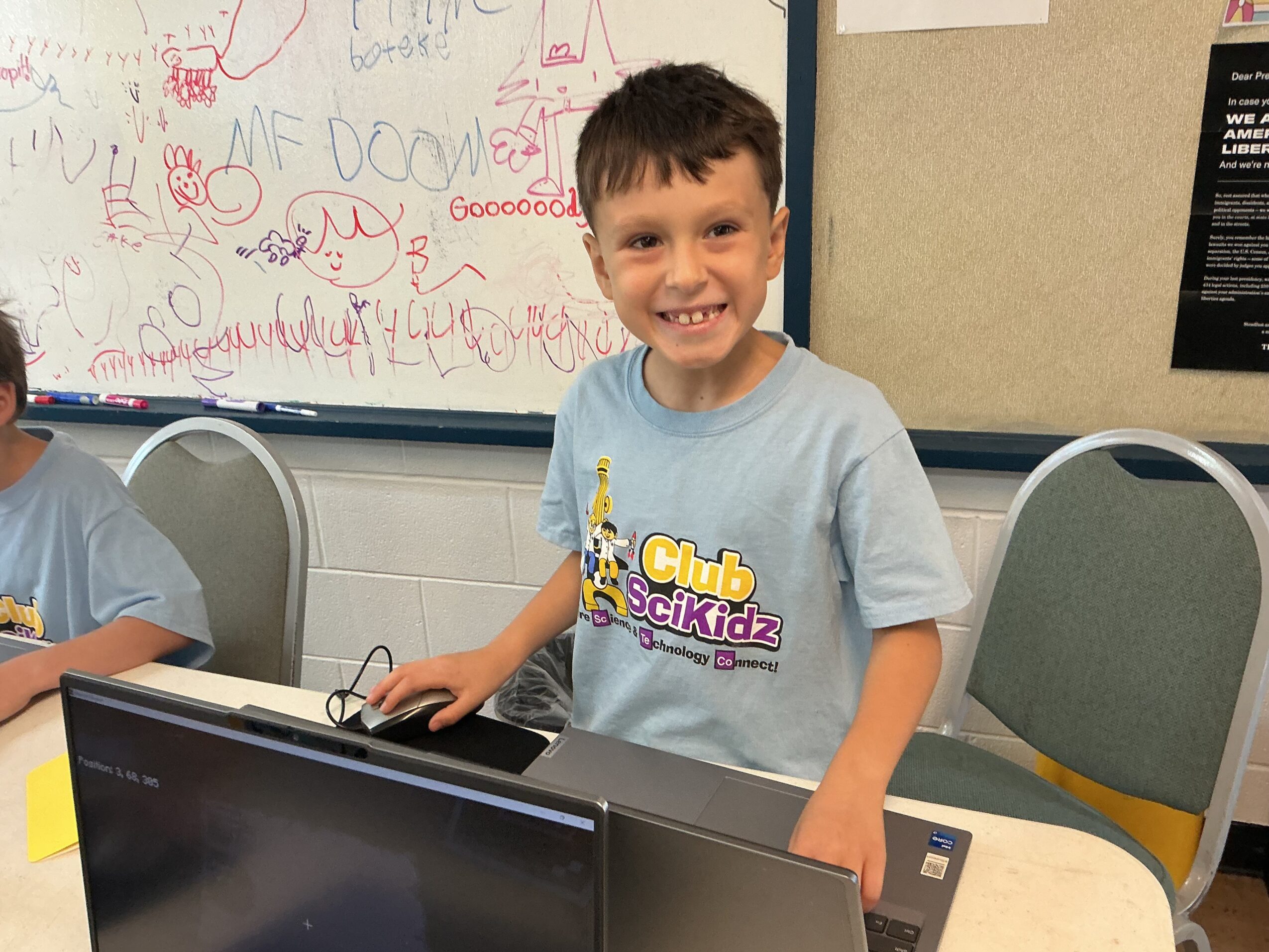 A smiling child stands beside a laptop in a classroom, with a decorated whiteboard and chairs behind him.