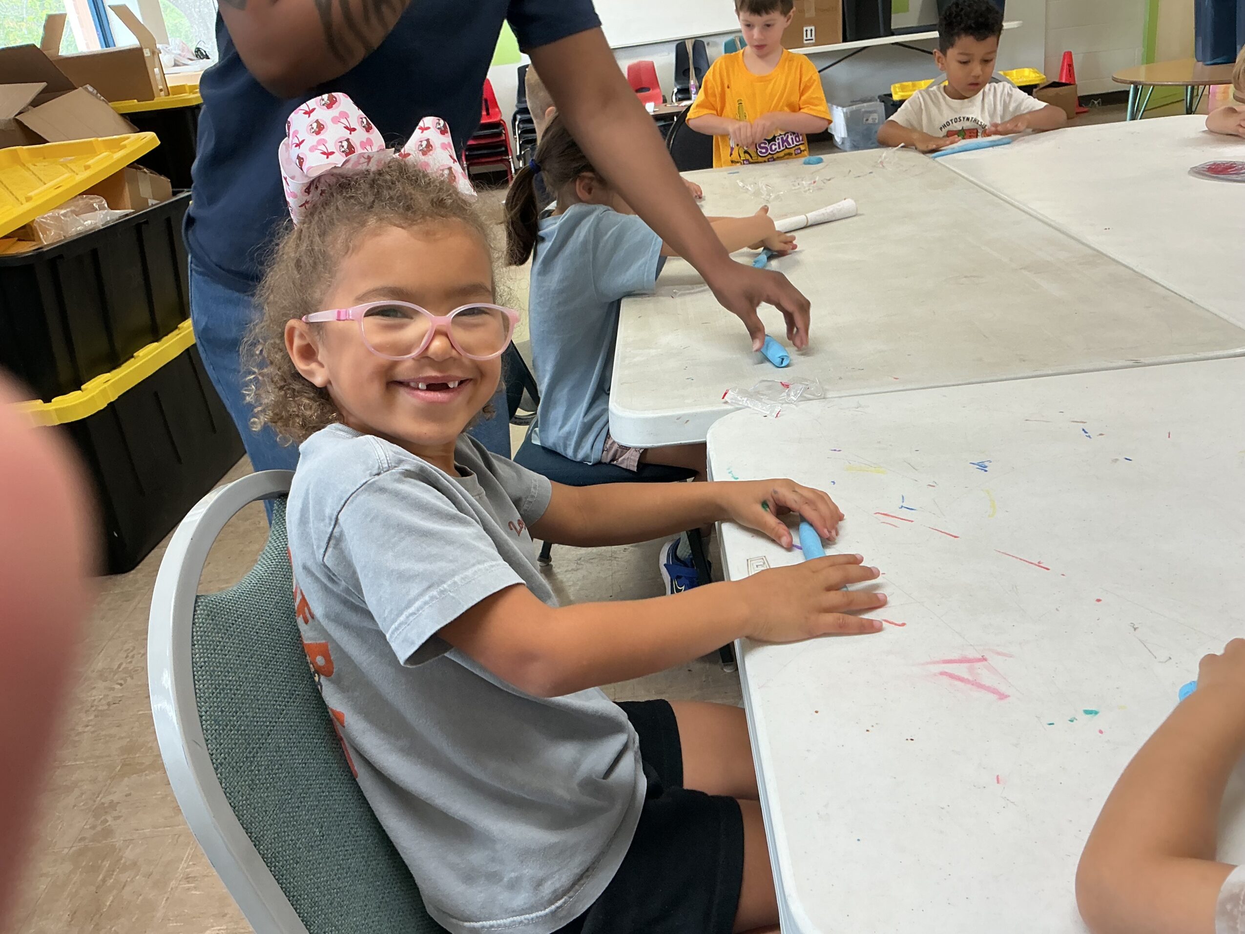 A child wearing glasses and a bow sits at a classroom table while children draw in the background.