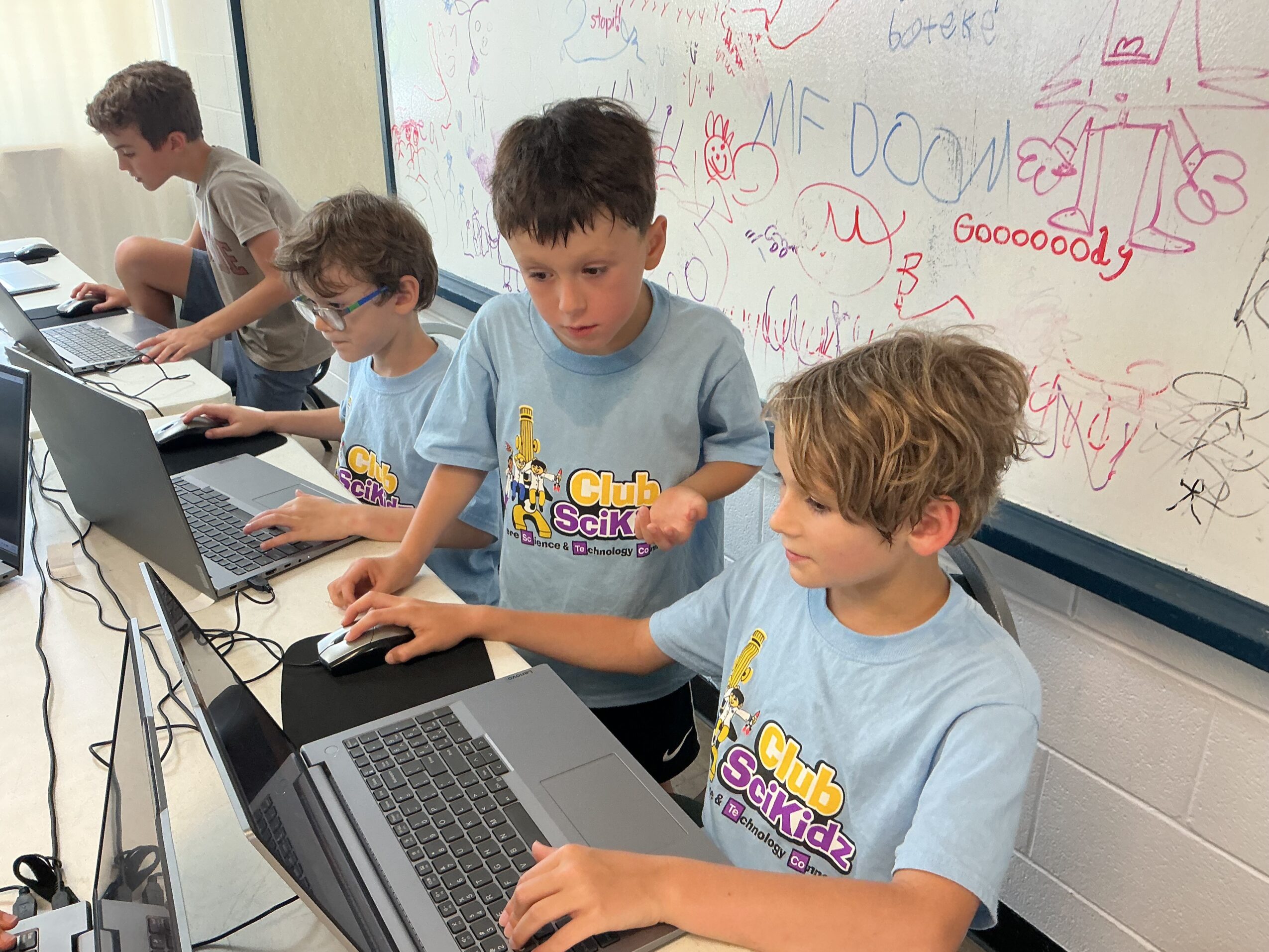 Four children work on laptops in front of a whiteboard covered with colorful drawings and writing.