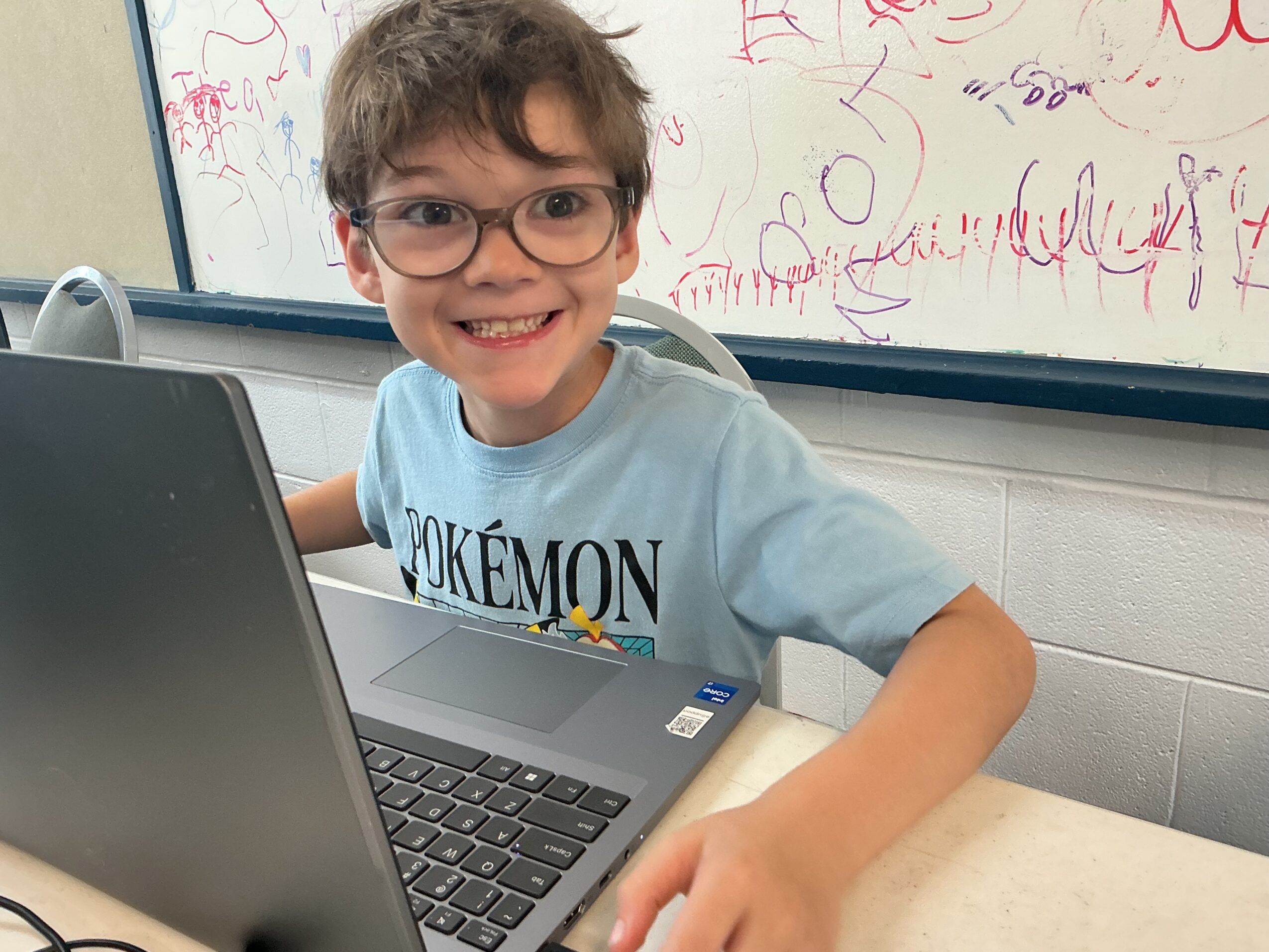 A smiling child with glasses sits at a laptop in front of a whiteboard covered with writing and drawings.