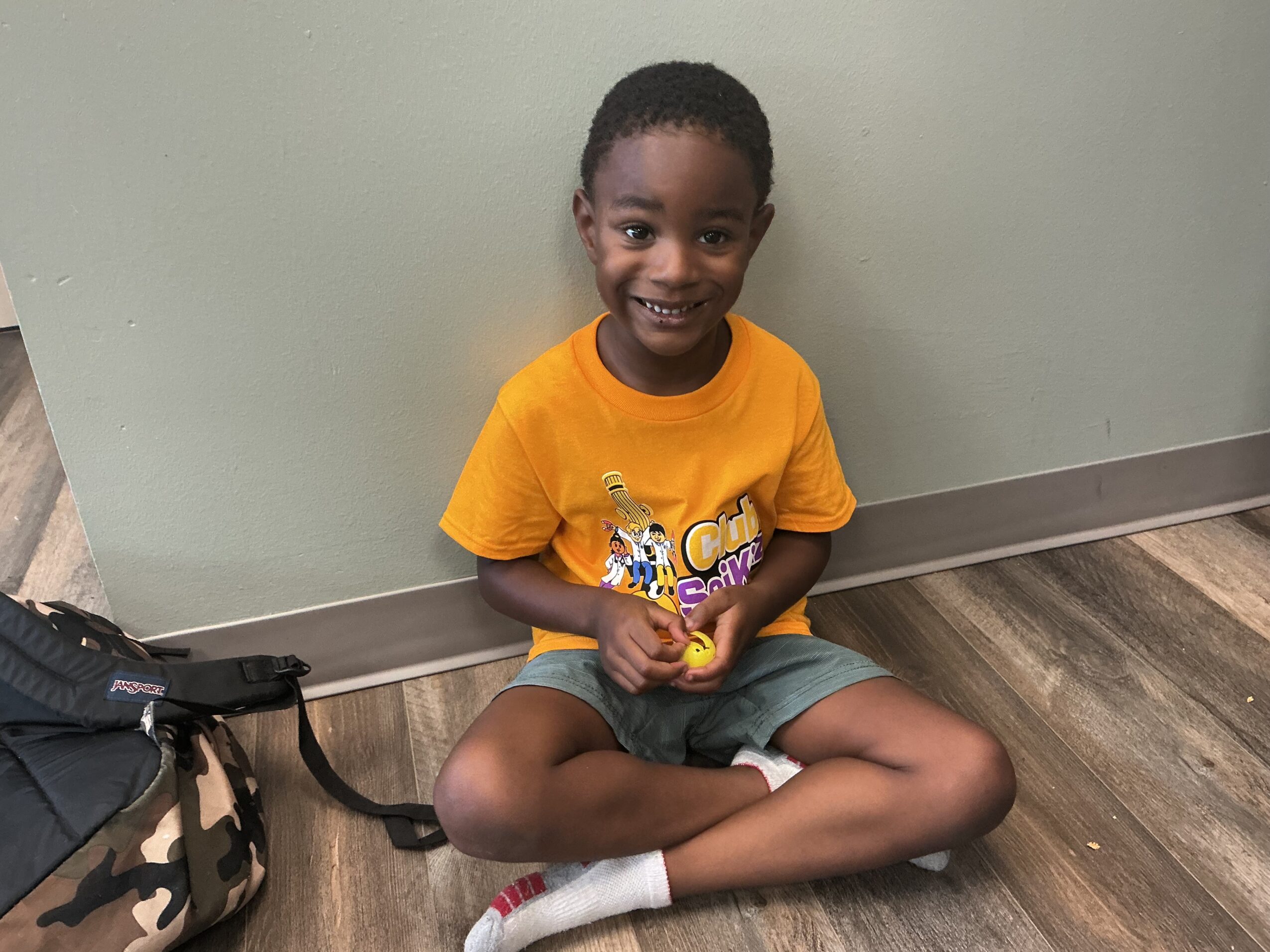 A child sits cross-legged on a wood-patterned floor, wearing an orange shirt and gray shorts beside a backpack.