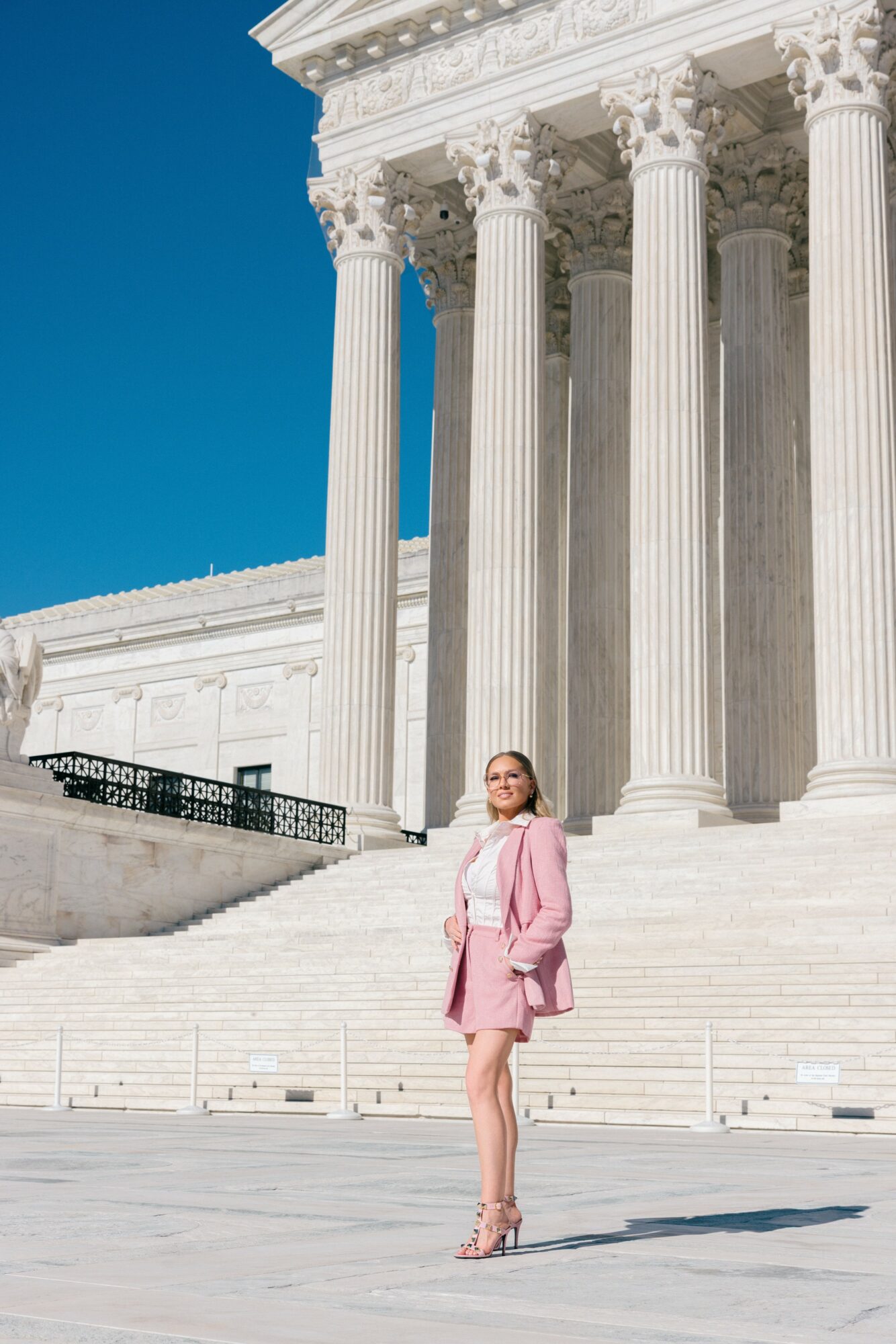 Woman in pink dress and heels standing on steps in front of large white classical building with columns, blue sky background.
