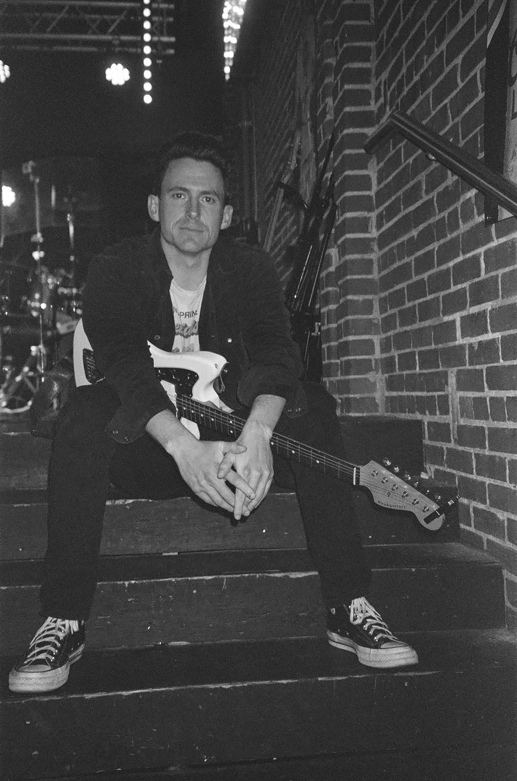 Person seated on steps holding an electric guitar beside a brick wall.