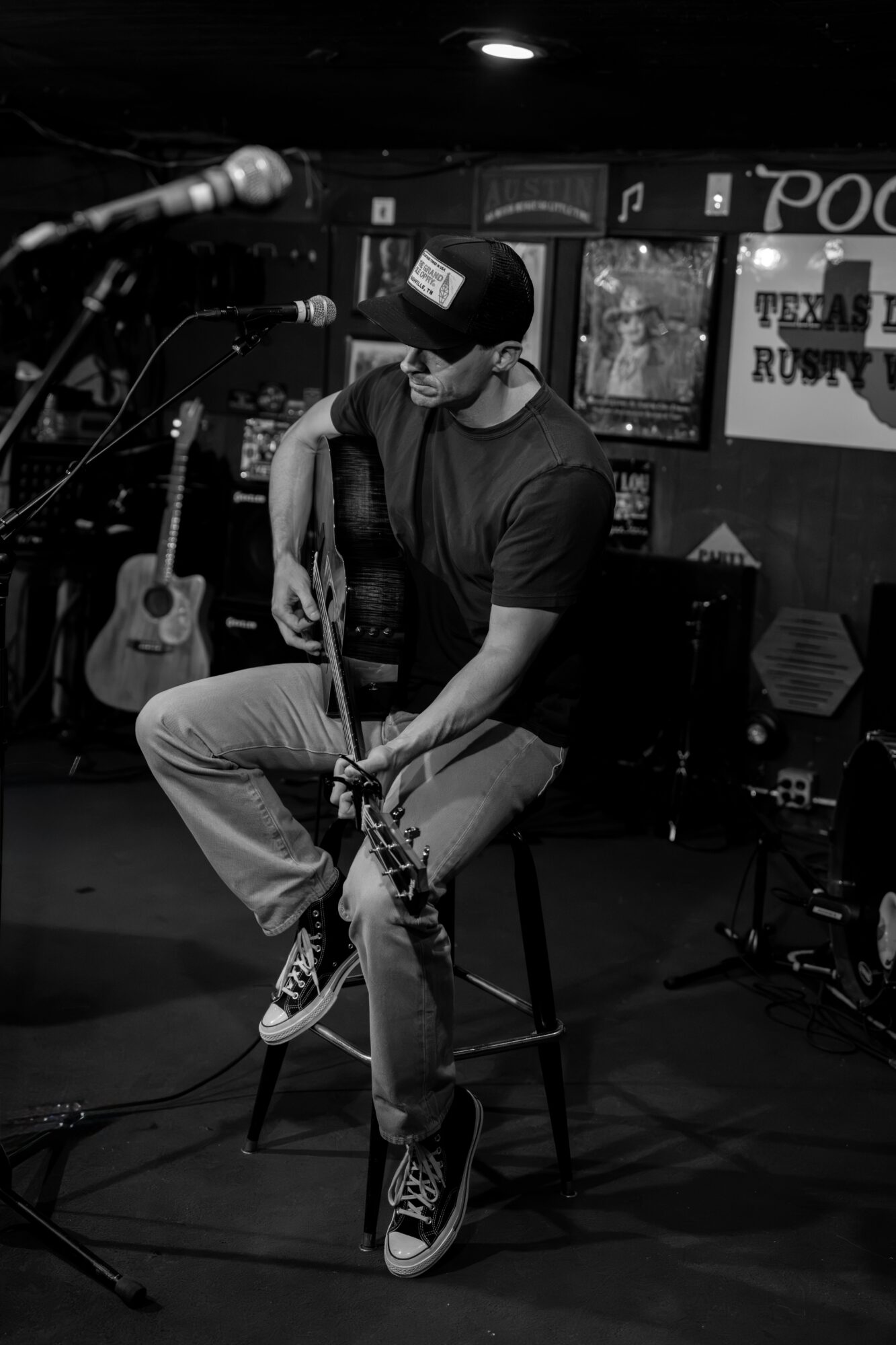 A seated person plays an acoustic guitar onstage beside a microphone, with posters and equipment in the background.
