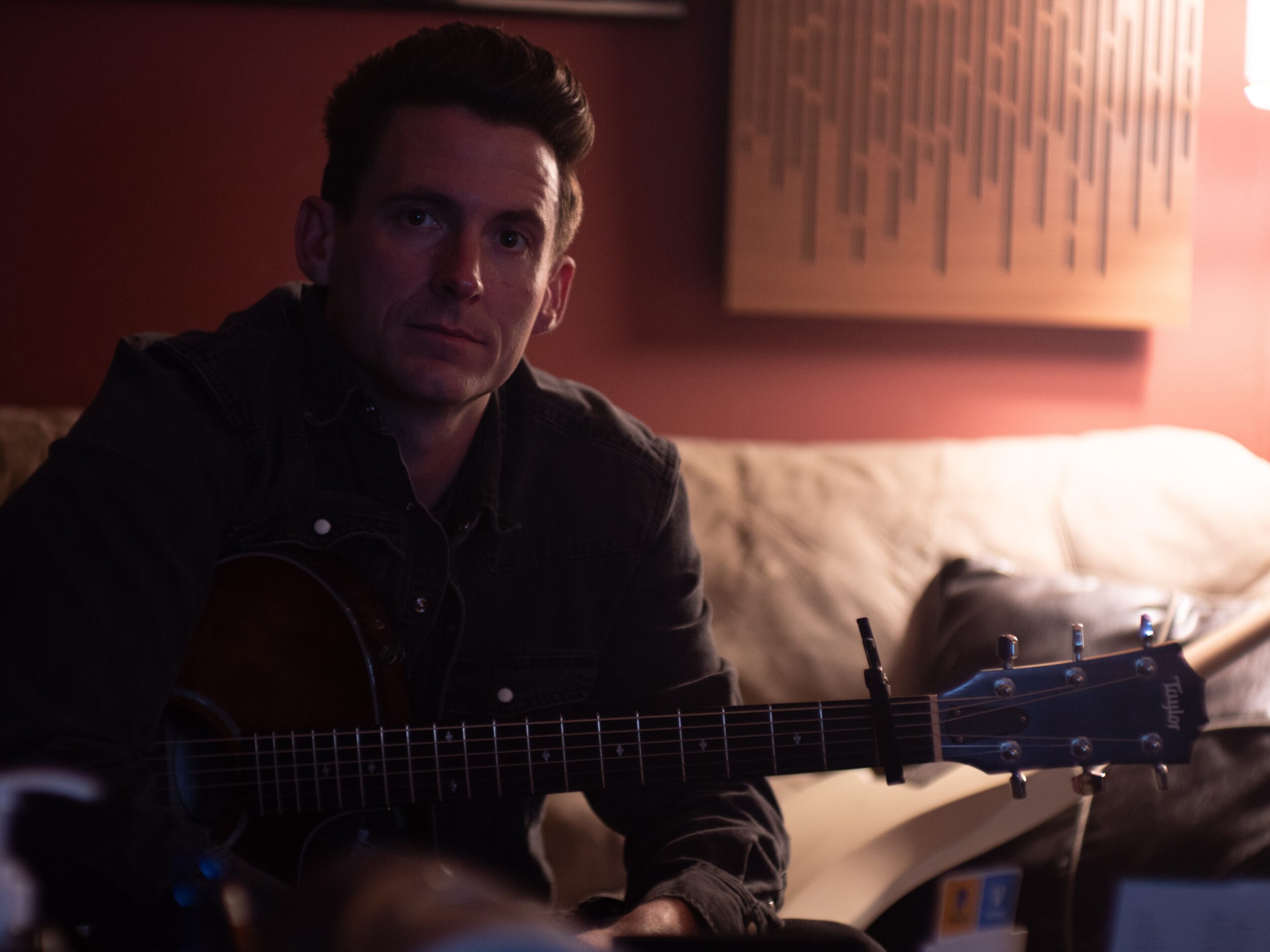 A seated person holds an acoustic guitar in a dimly lit room.