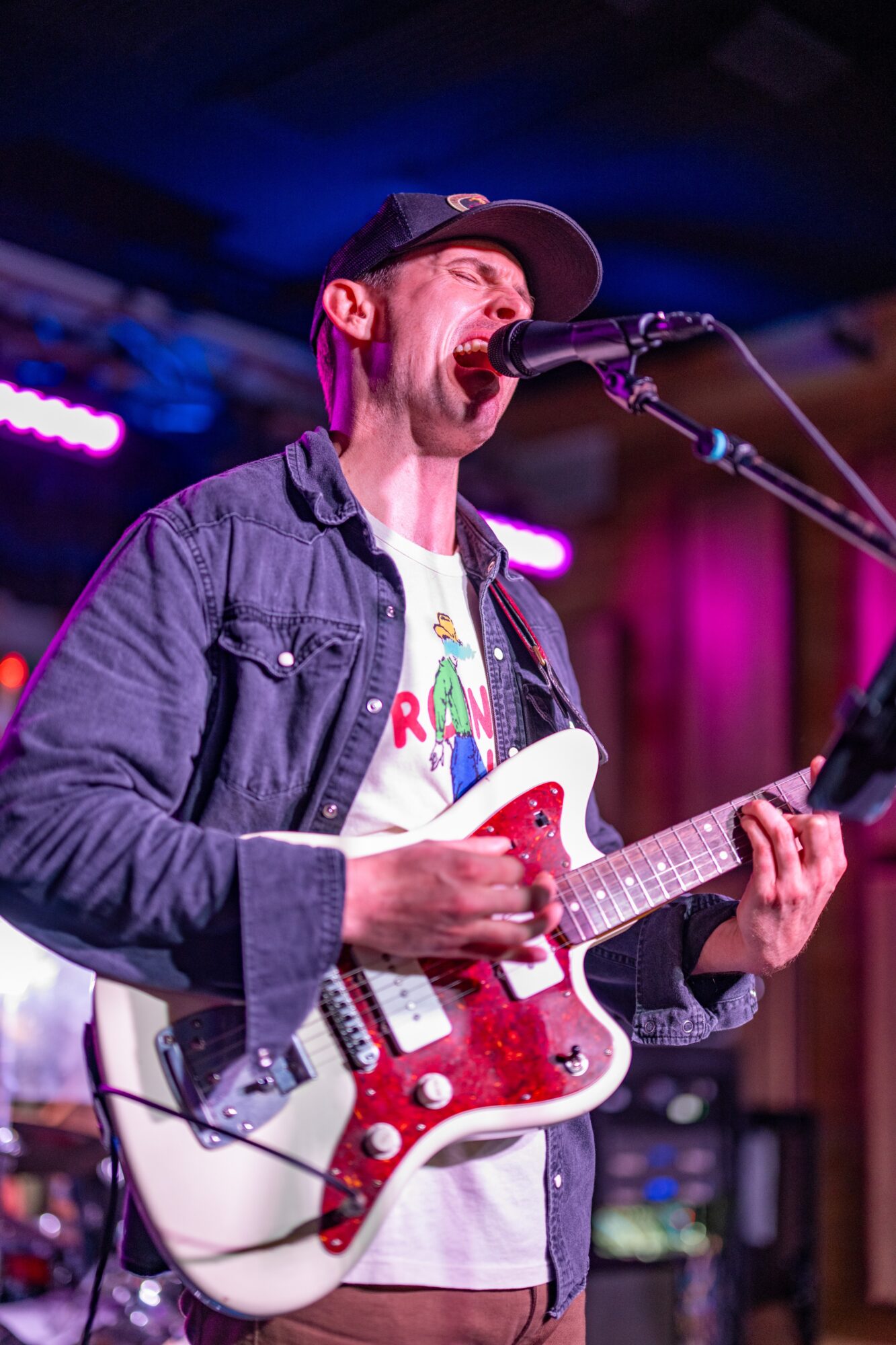 A person in a cap sings into a microphone while playing a red-and-white electric guitar onstage.