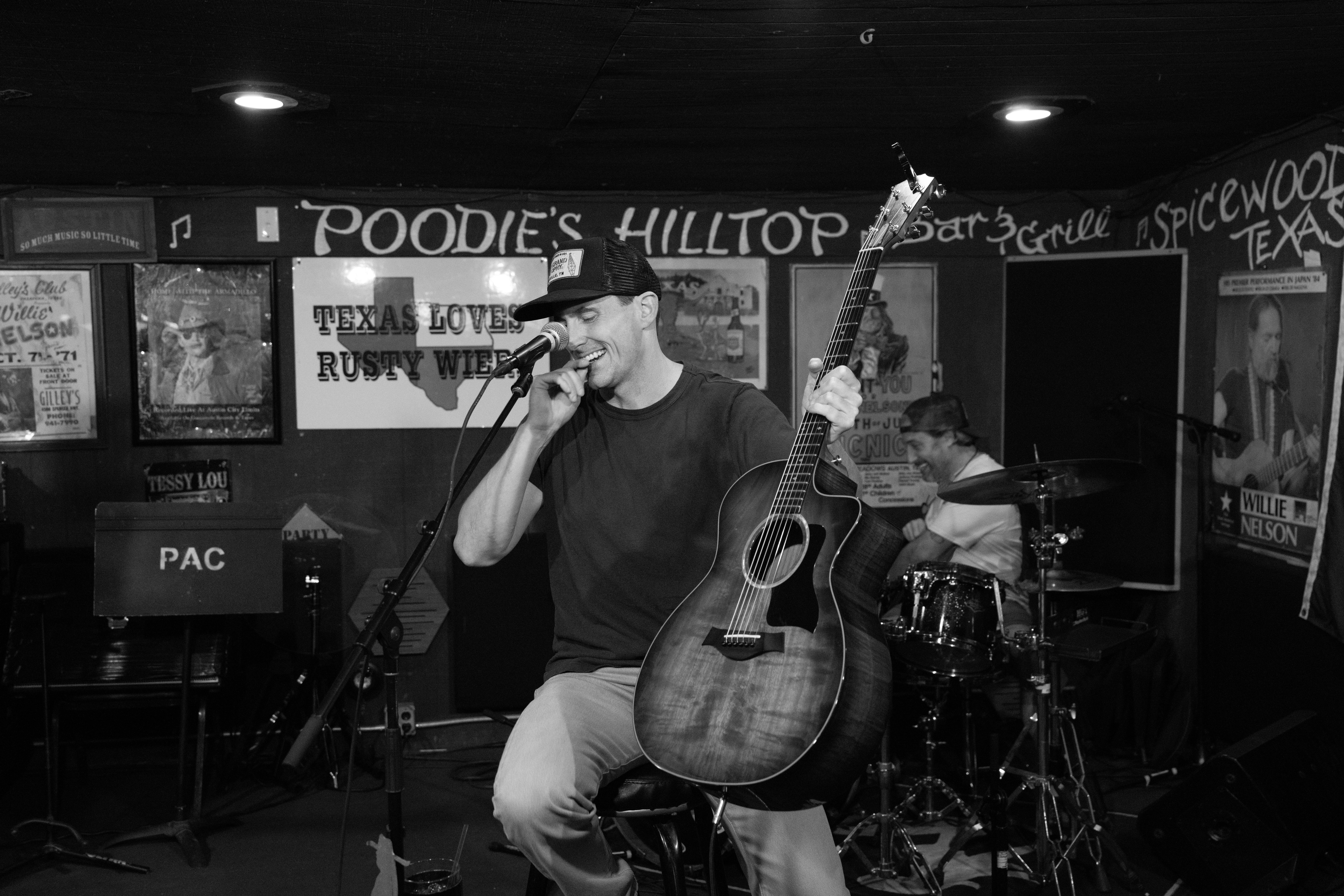 A musician sings into a microphone while playing guitar on a small stage, with a drummer behind him.