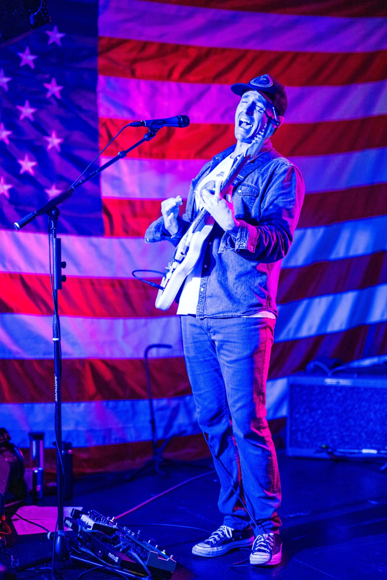 A musician plays guitar beside a microphone in front of a large American flag backdrop.