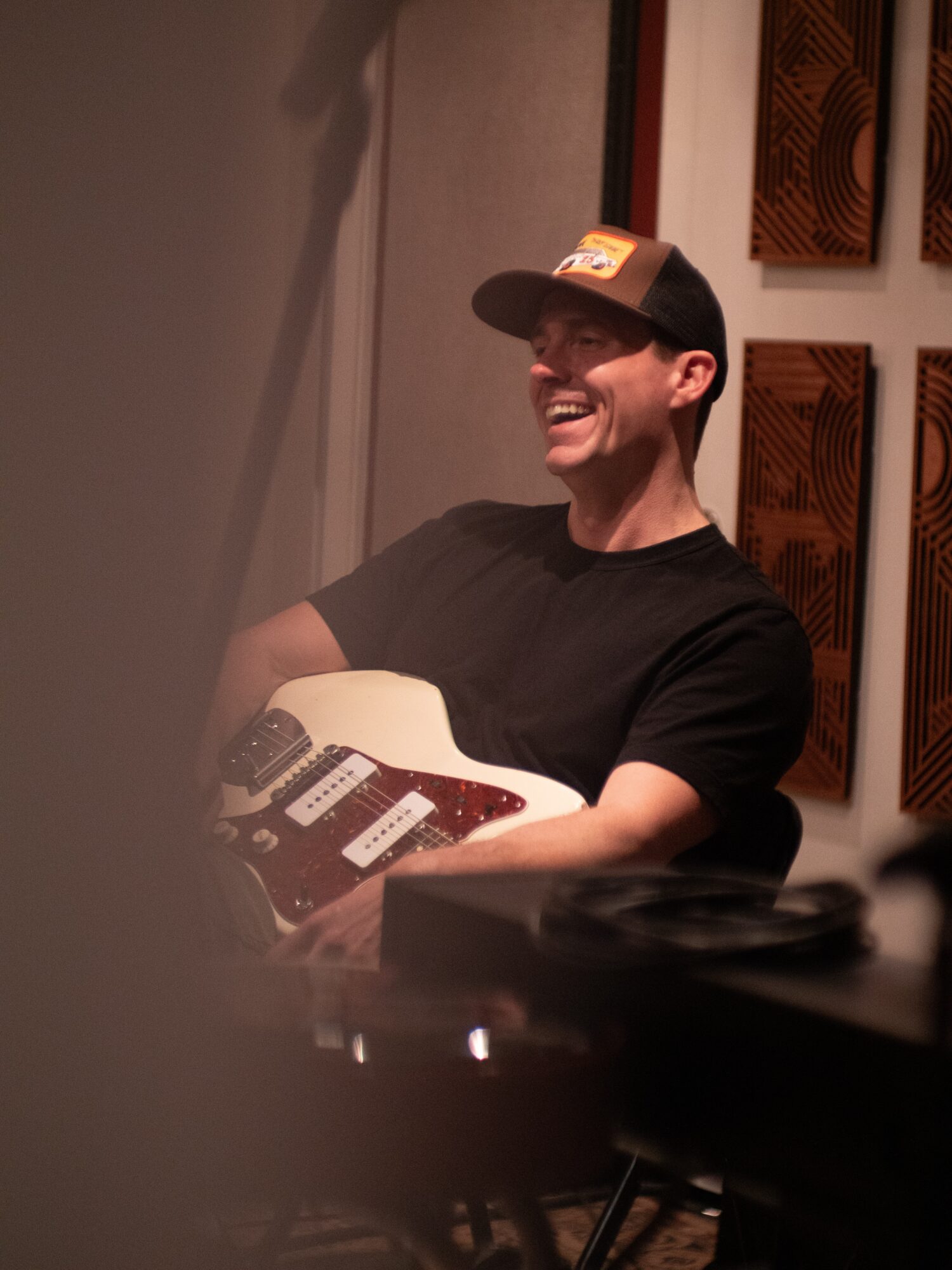 A seated person wearing a cap holds an electric guitar in a dim room with framed wall art.