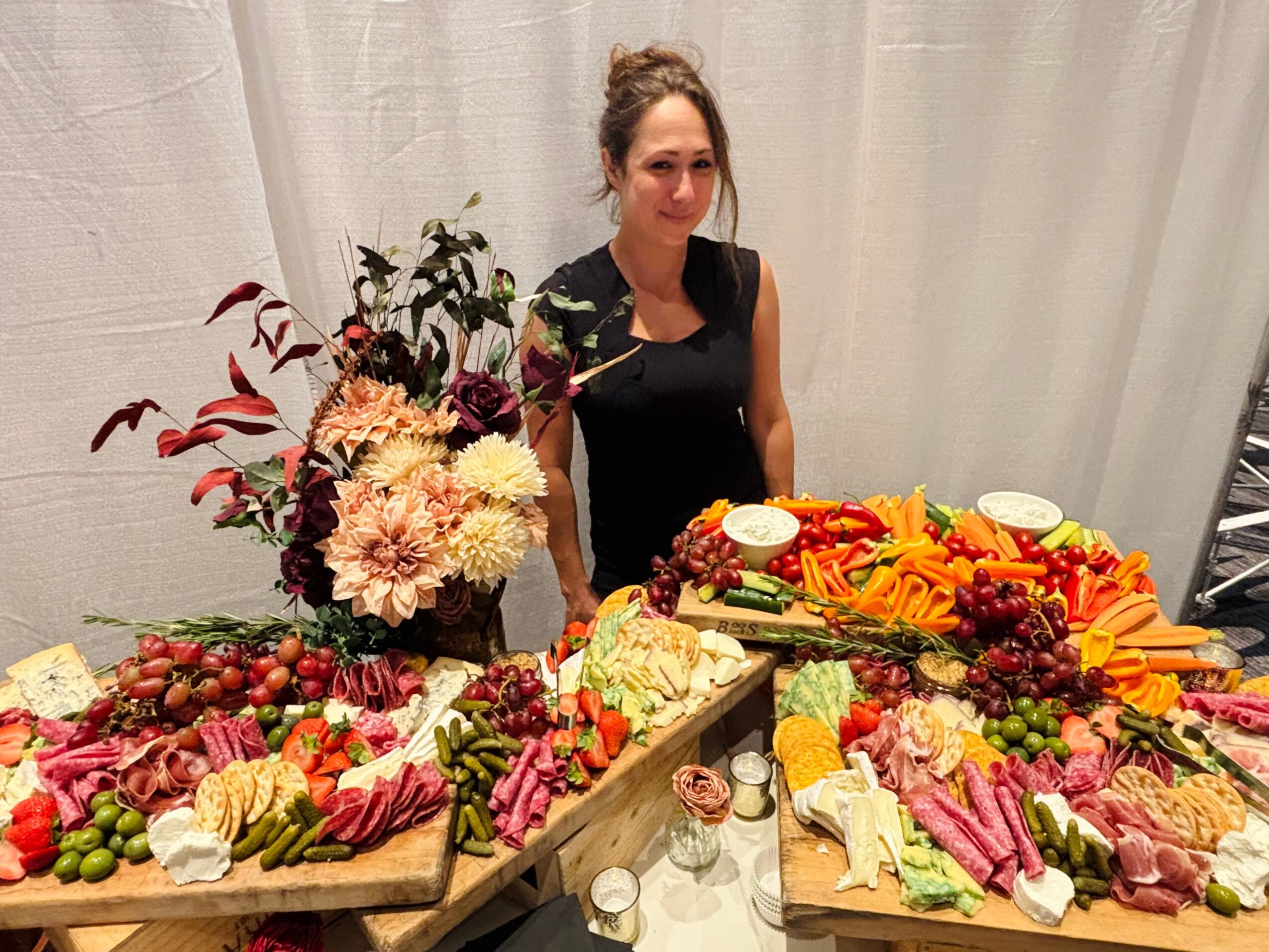 A person stands behind a table laden with flowers, fruits, vegetables, bread, and assorted foods.