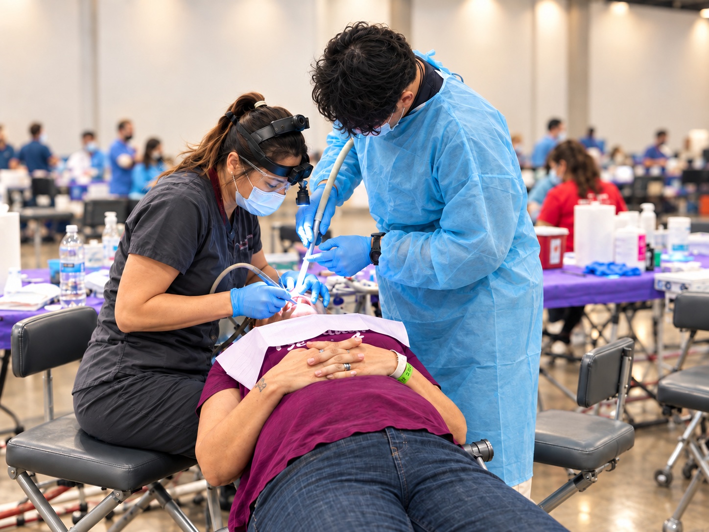 Two masked healthcare workers treat a person lying on a chair in a large indoor clinic.