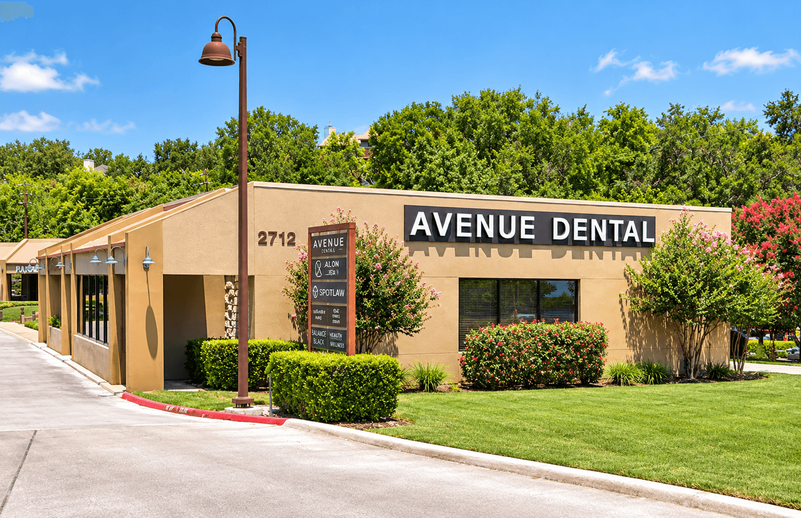 Single-story dental office with a sign reading “Avenue Dental,” landscaped shrubs, lawn, and a parking area.