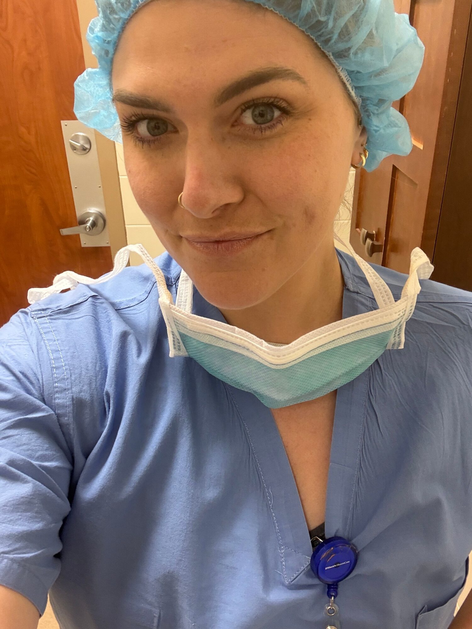 A person in blue medical scrubs and a hair cap, with a surgical mask hanging below their chin.