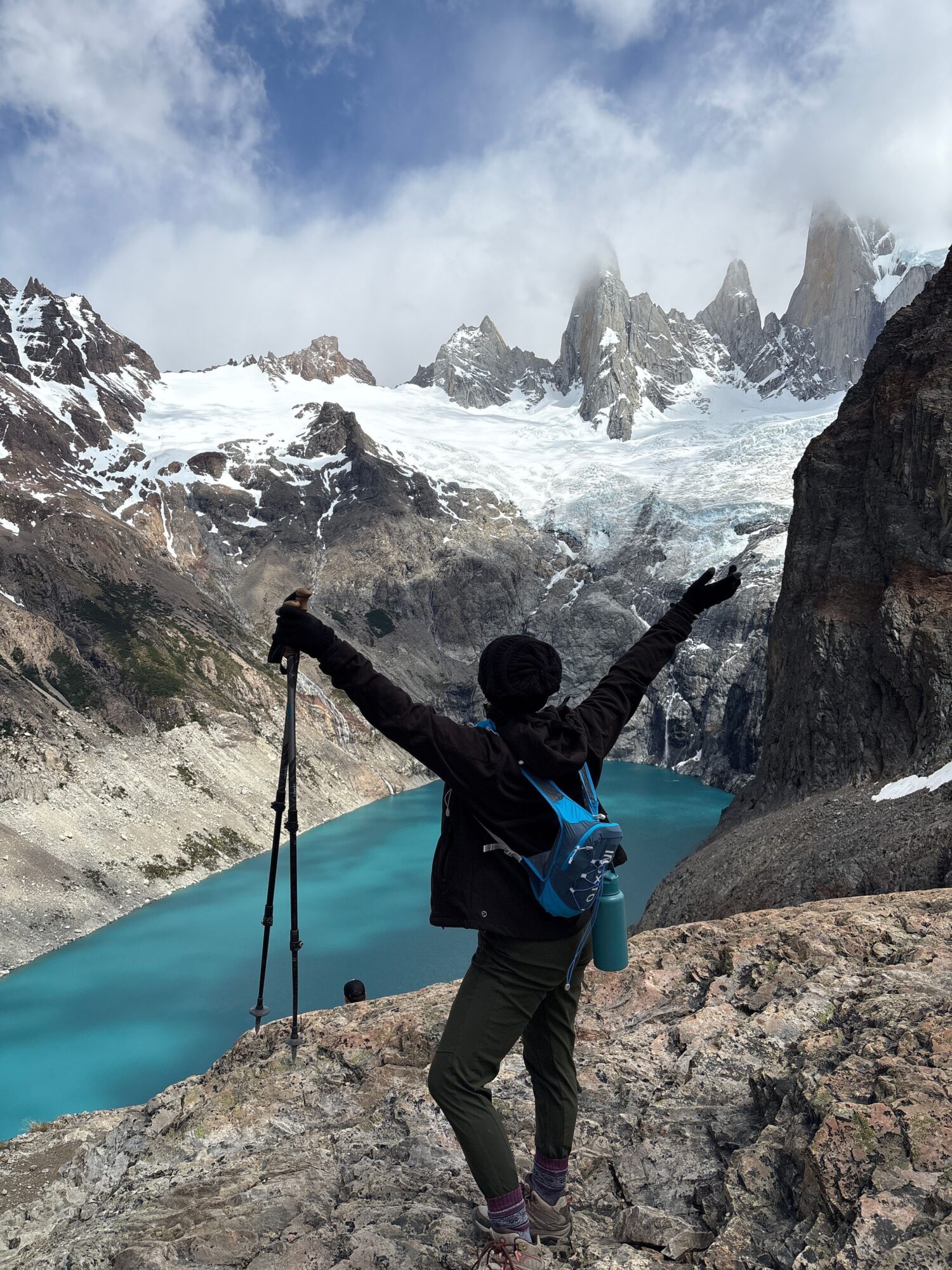 A hiker stands on a rocky ledge above a turquoise mountain lake, with snow-covered peaks and clouds beyond.