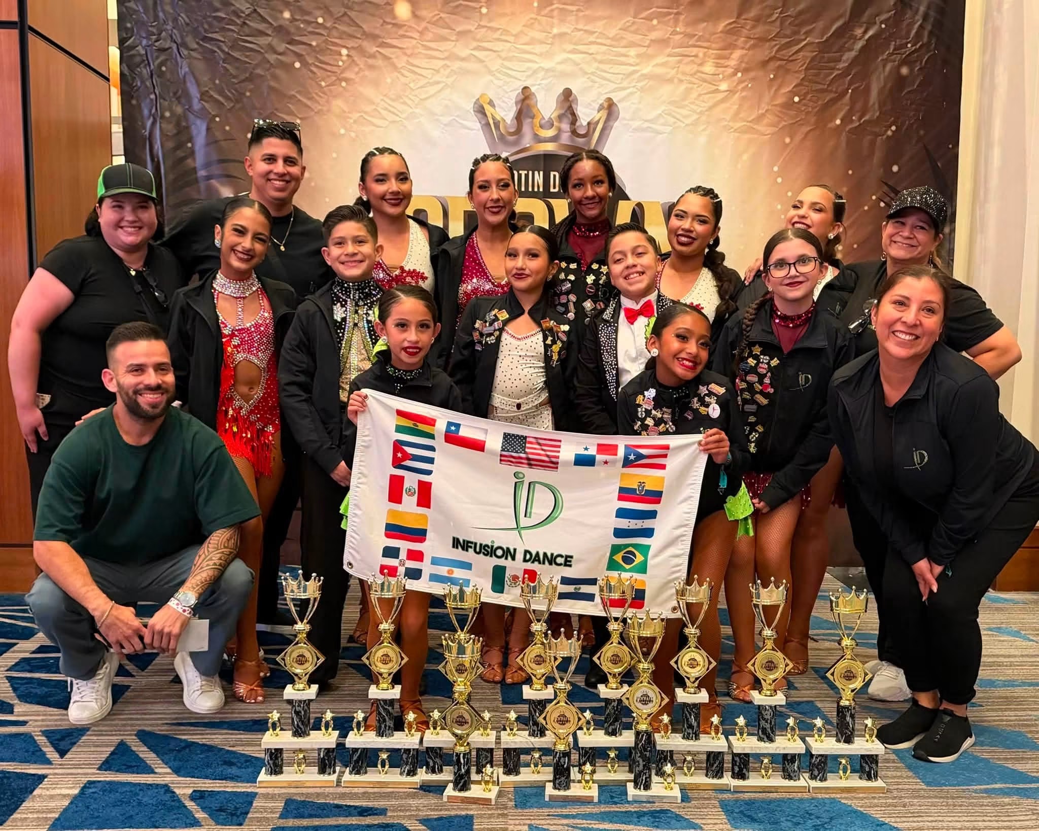 A group poses behind a banner and rows of dance trophies at an indoor event.